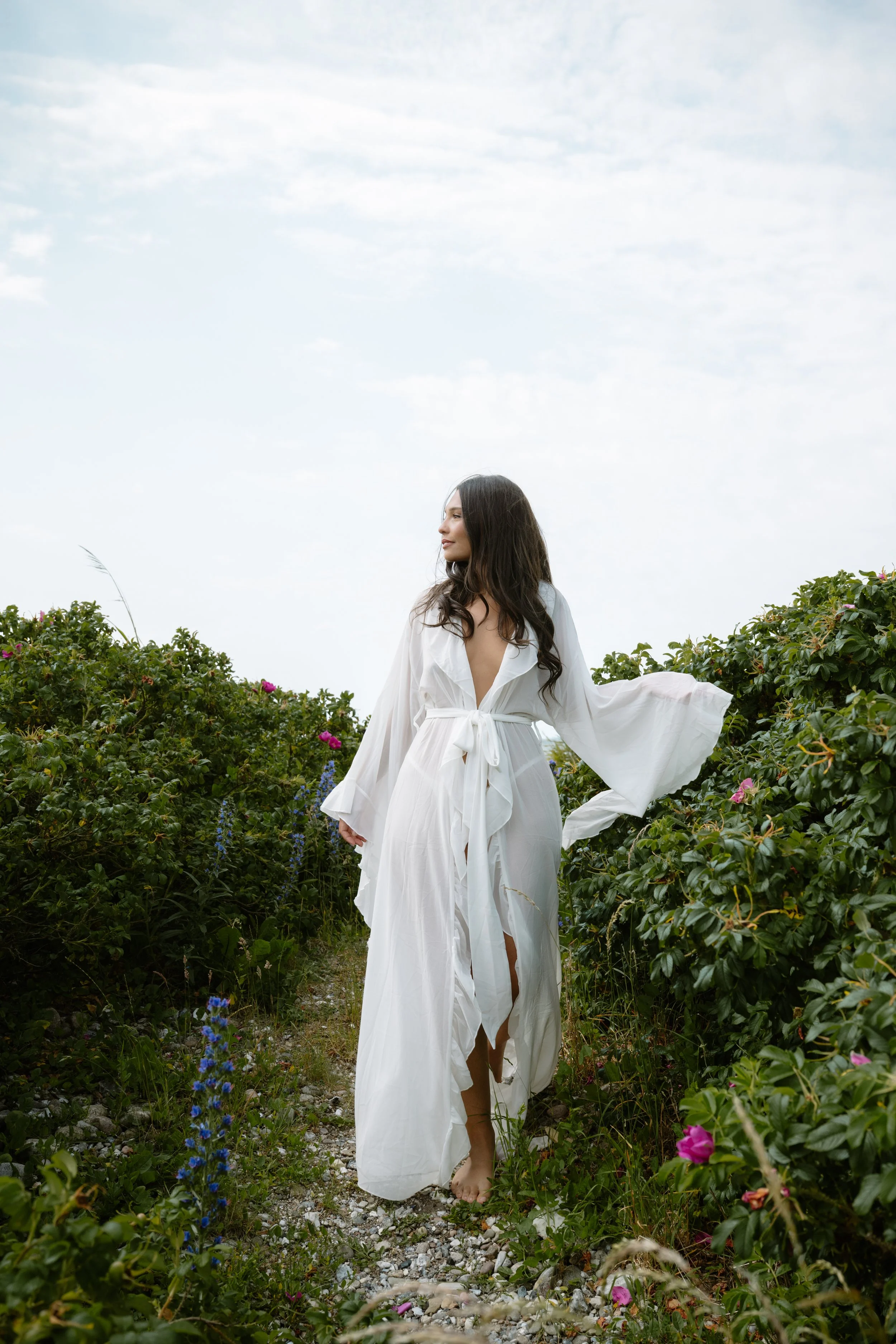 A woman wearing a long white dress walking through a lush, flower-filled path outdoors under a cloudy sky.