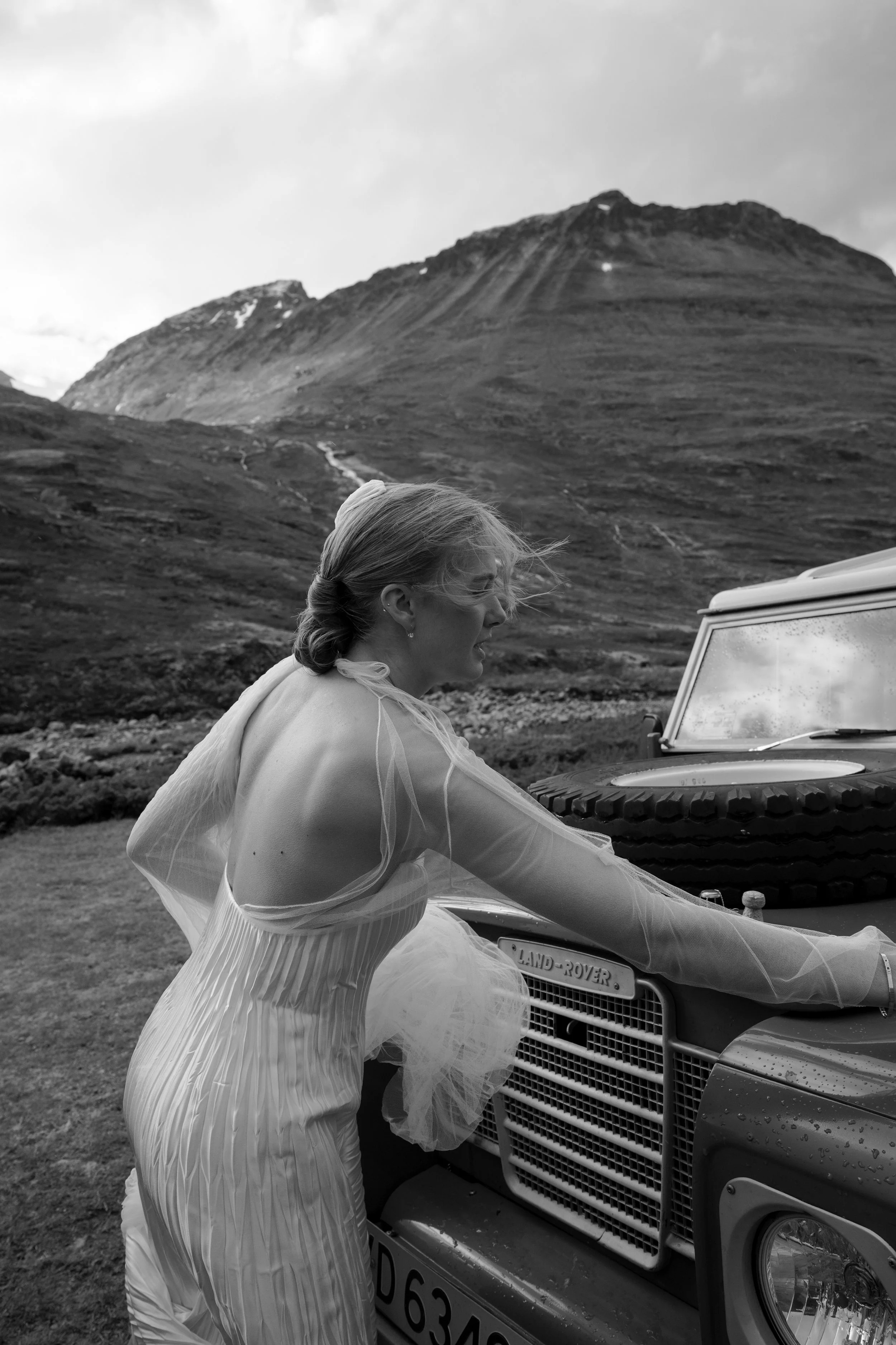 A woman in a wedding dress leaning on a Land Rover vehicle outdoors with mountains in the background.