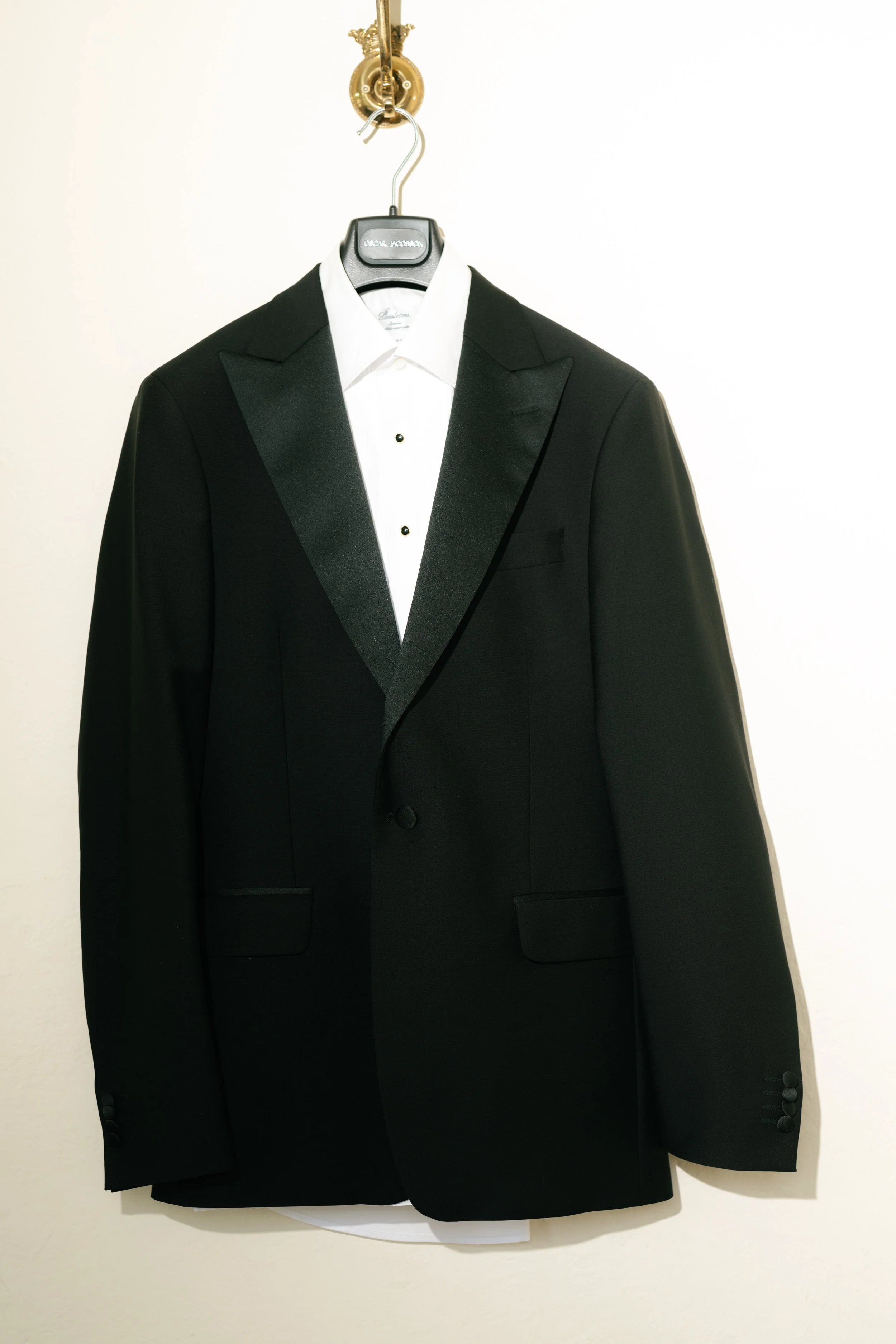Black tuxedo jacket with satin peaked lapels over a white dress shirt with black buttons, hanging on a hanger against a plain white wall.