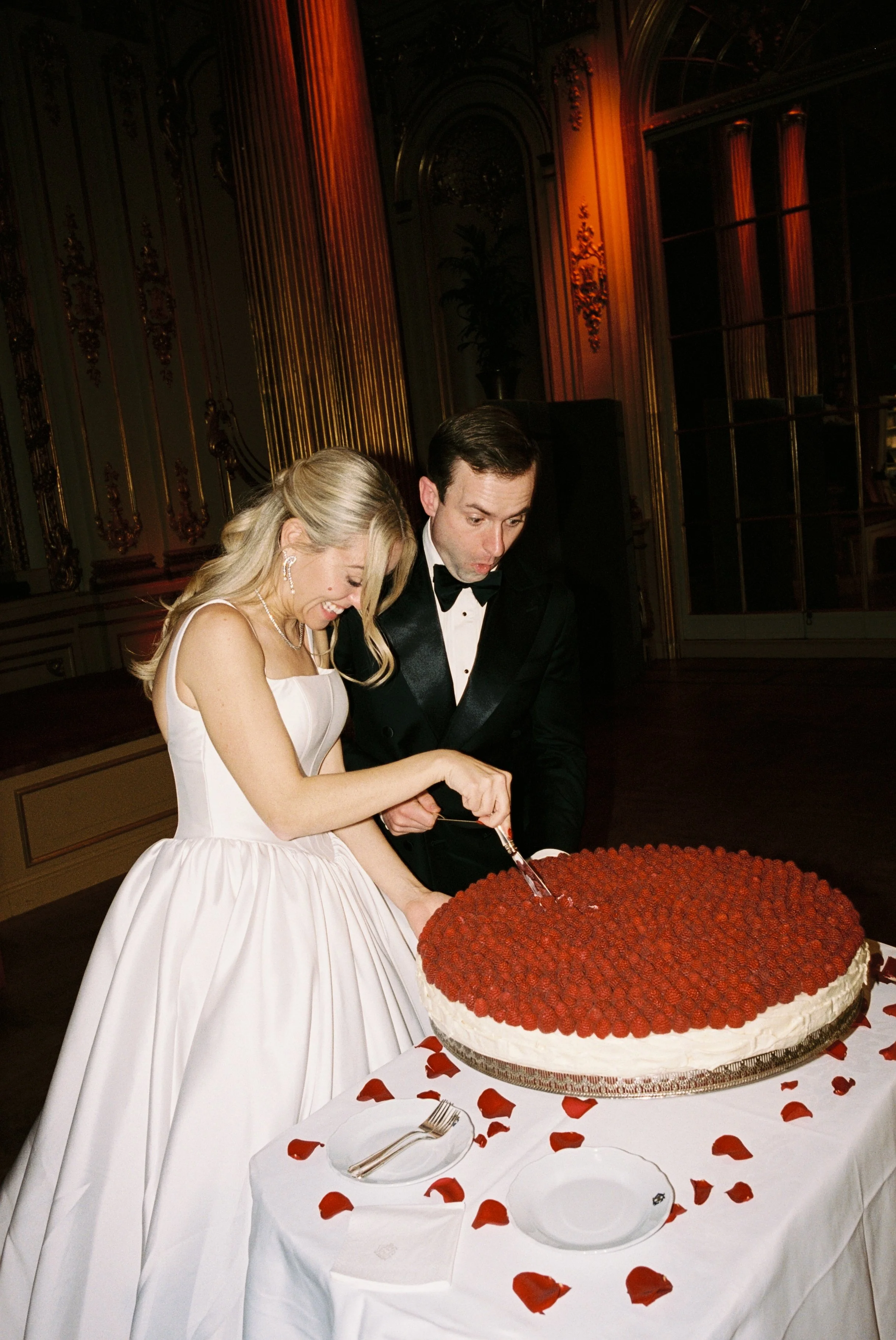 A bride and groom cut a large wedding cake decorated with strawberries, surrounded by rose petals, in an elegant, ornate room.