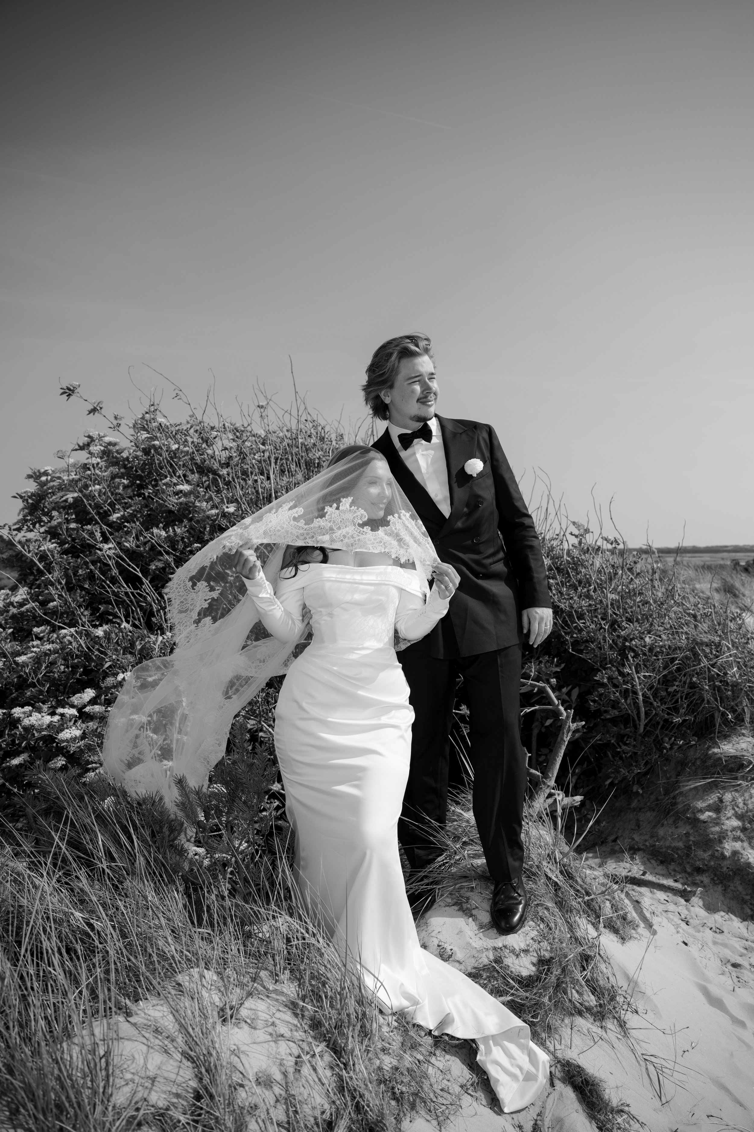 Black and white photo of a bride and groom standing outdoors on a sandy area with bushes behind them. The bride is wearing a strapless wedding dress and veil, while the groom is in a tuxedo with a bow tie and boutonniere. The bride is holding her vei