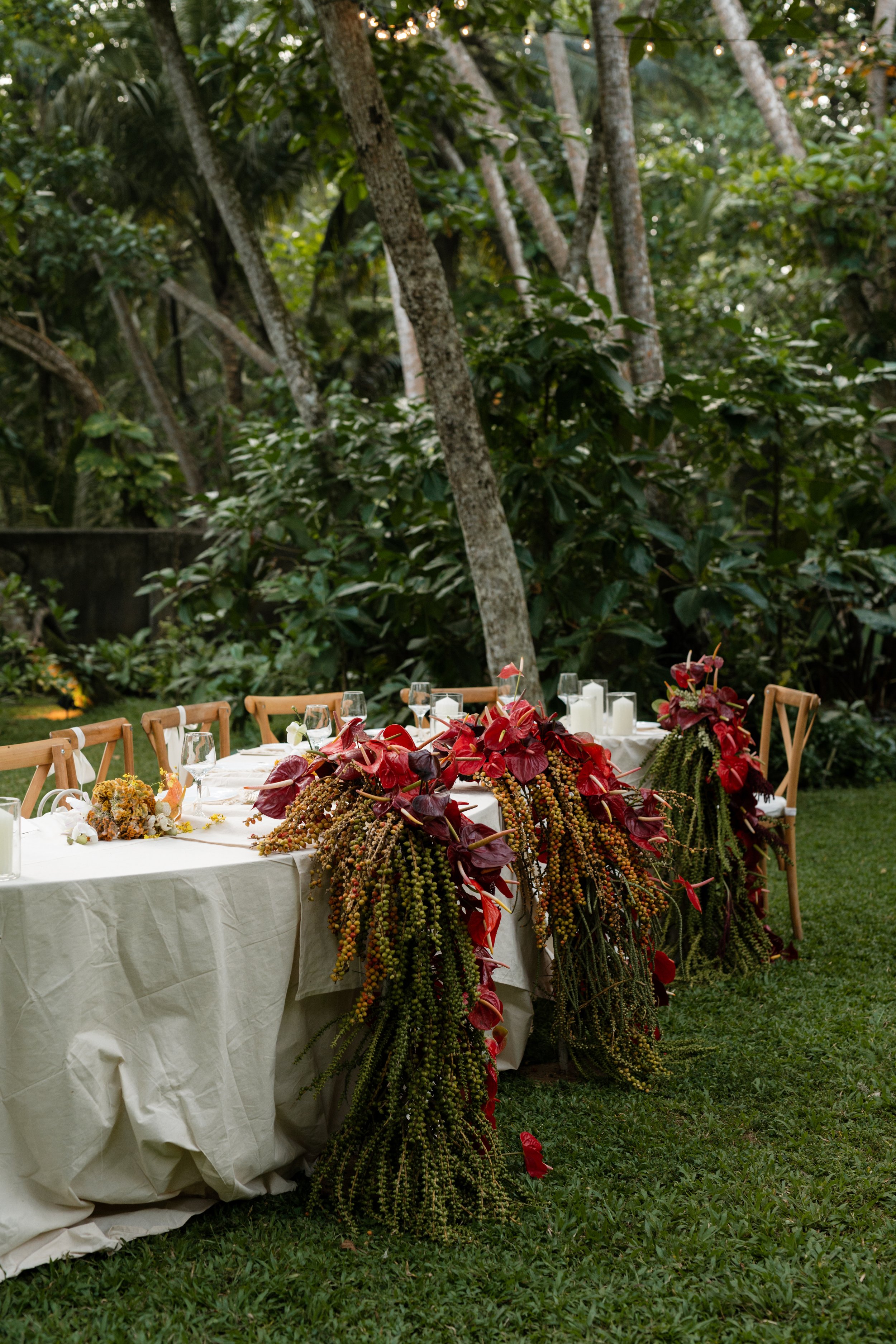 Outdoor dining table set on grass, with elegant floral arrangements featuring red and burgundy flowers and greenery, surrounded by wooden chairs, in a lush garden setting