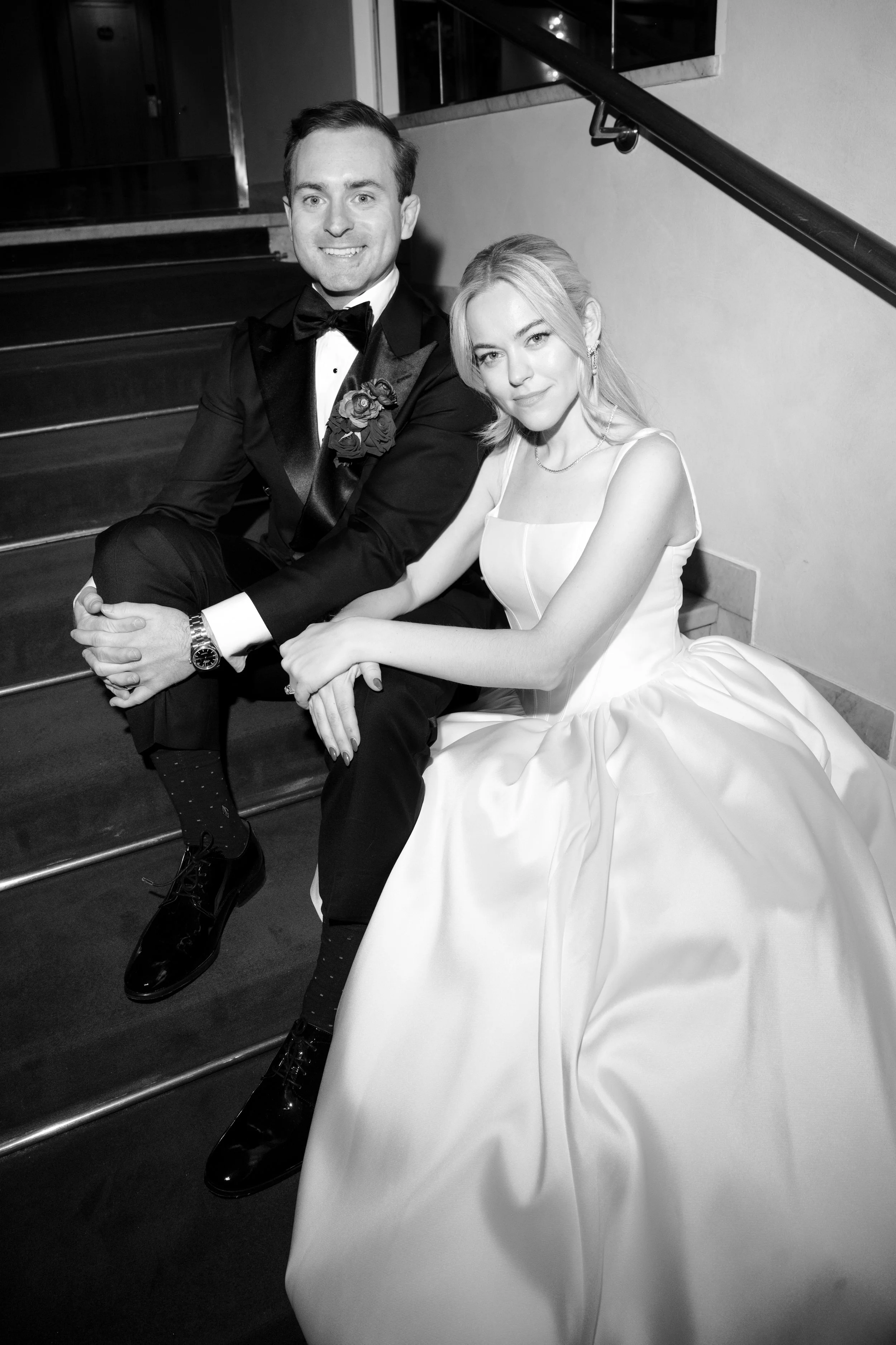A black-and-white photo of a couple in formal attire sitting on stairs, smiling at the camera. The man is wearing a tuxedo with a bow tie, and the woman is in a wedding gown. They appear to be newlyweds.