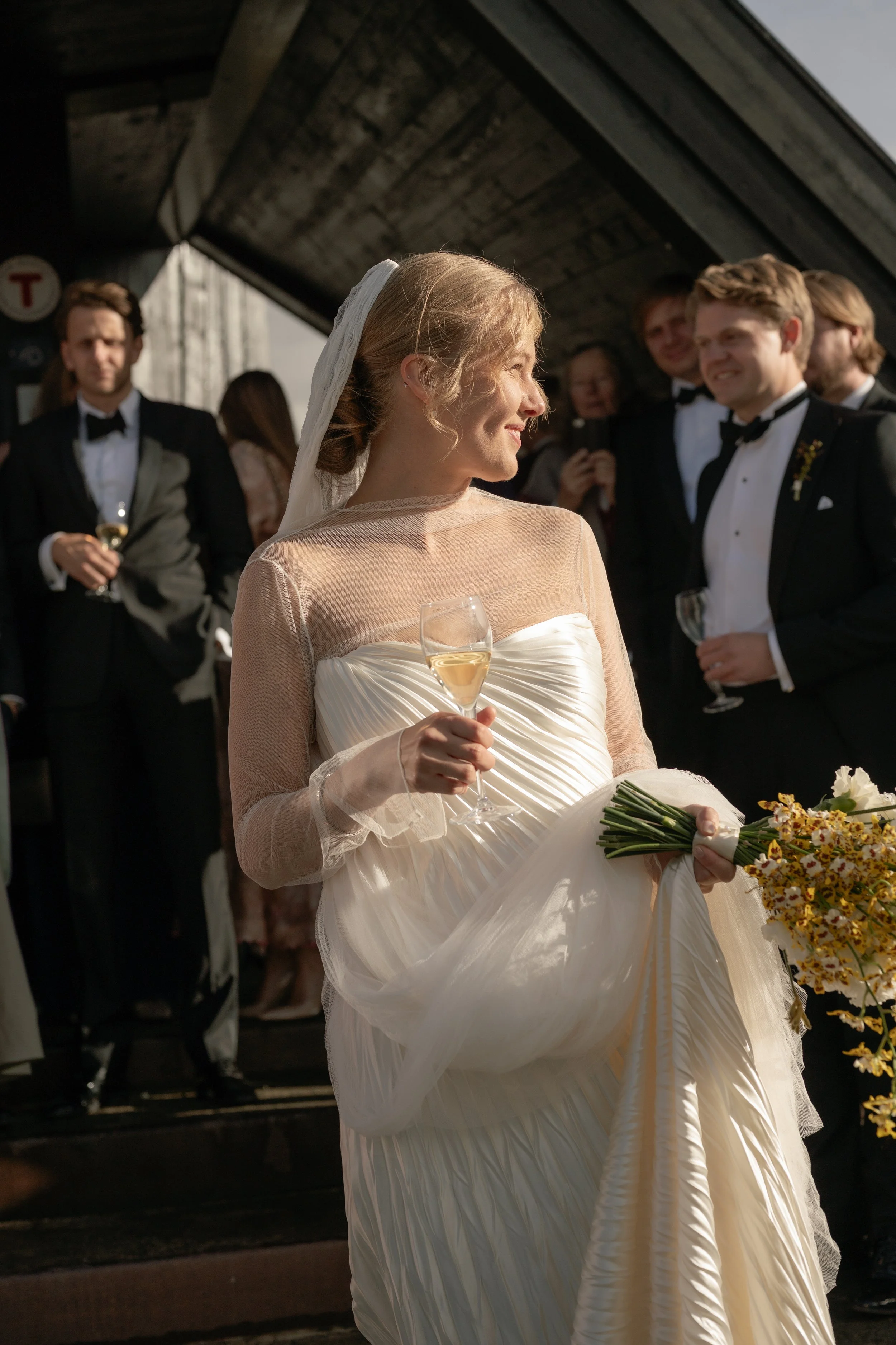 A bride in a white wedding dress and veil holding a glass of champagne and a bouquet of flowers, smiling and looking to her right, surrounded by groomsmen in tuxedos at a wedding celebration.