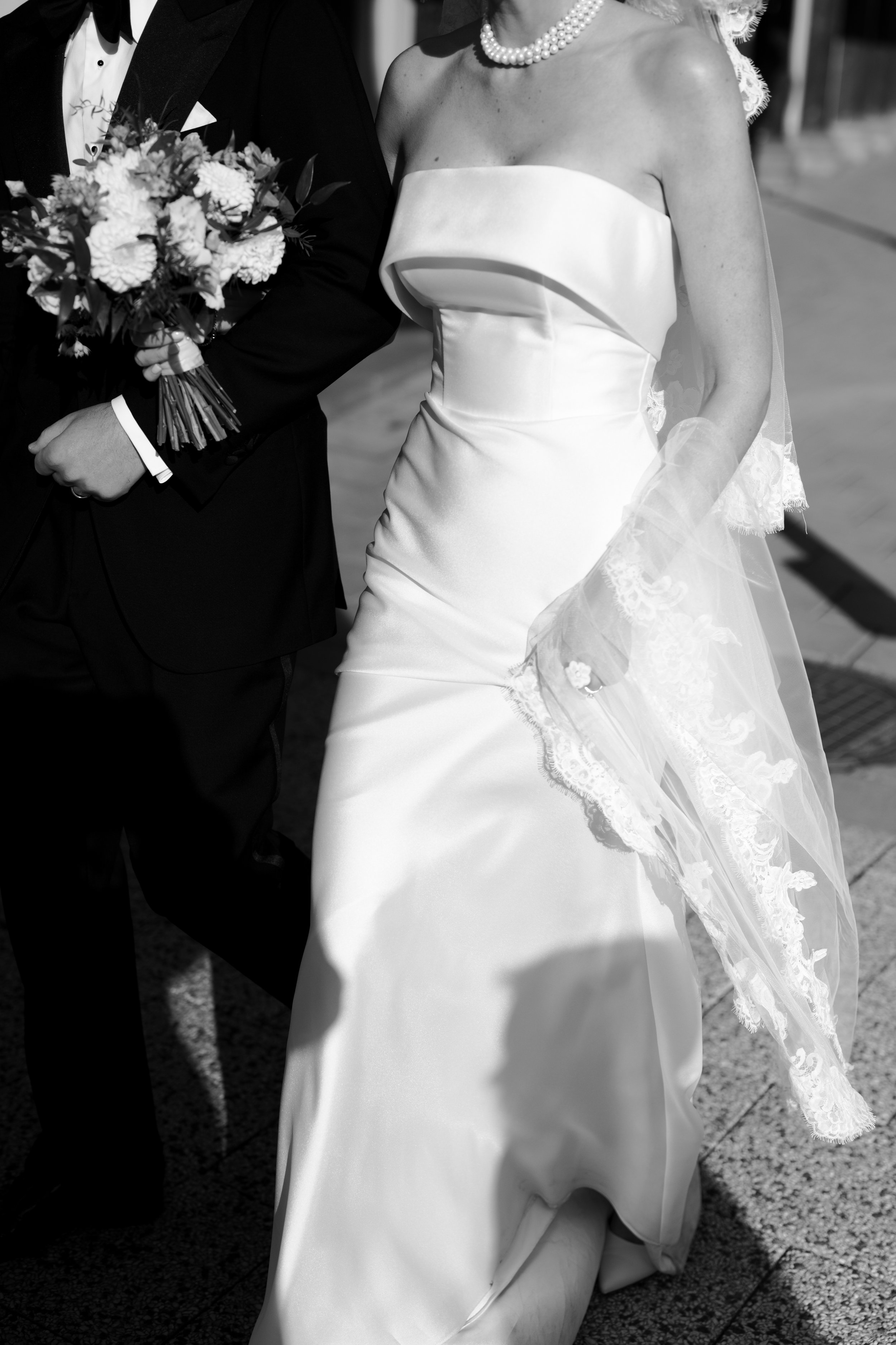 Black and white photo of a bride in a strapless wedding gown holding hands with a groom in a tuxedo, holding a bouquet of flowers.