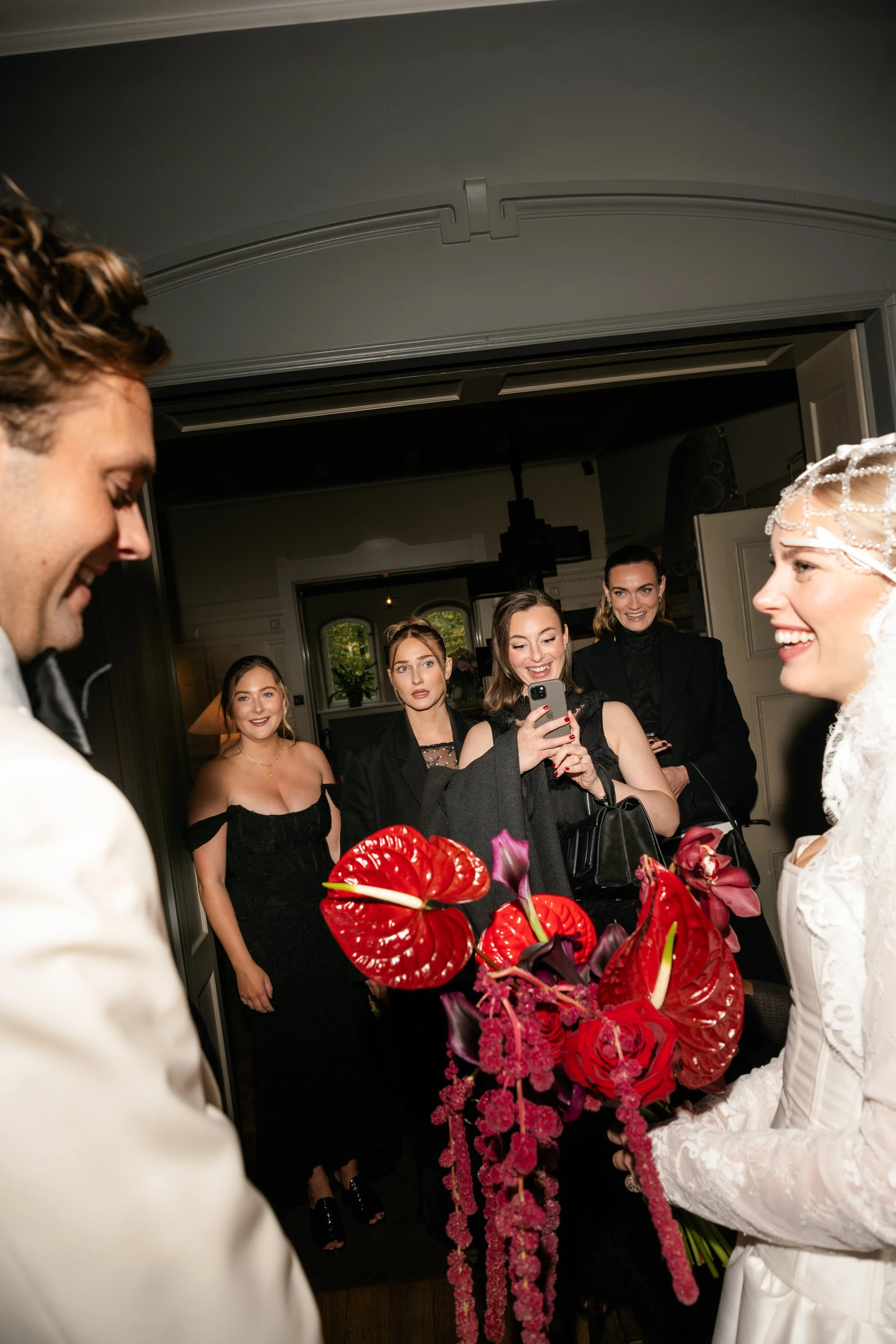 A bride holding a bouquet of red and purple flowers, smiling at the groom during a wedding celebration, with four women in black dresses and jackets smiling and taking pictures of her.