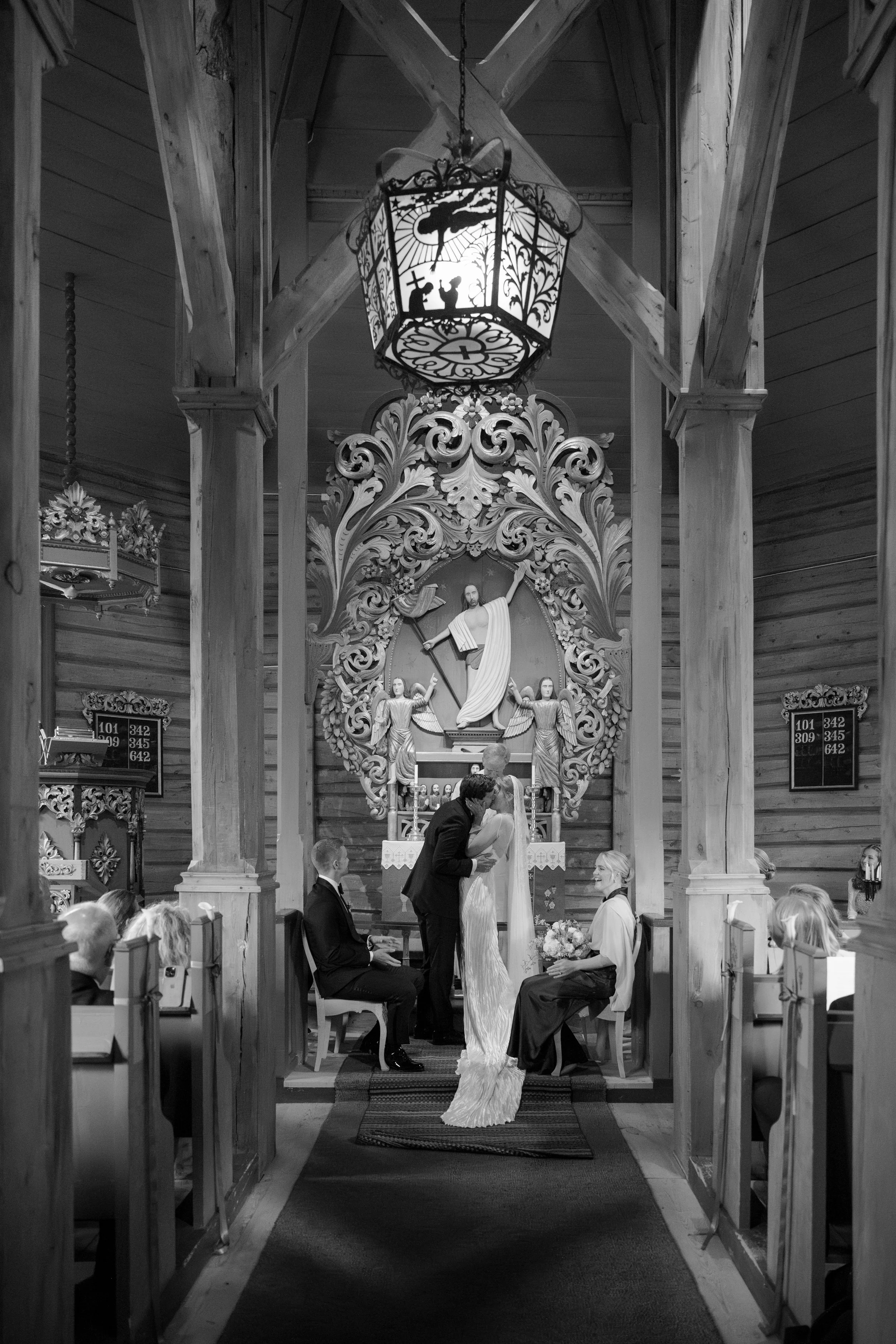 A black and white photo of a wedding ceremony inside a church, with a couple kissing at the altar, surrounded by witnesses and traditional religious decor.