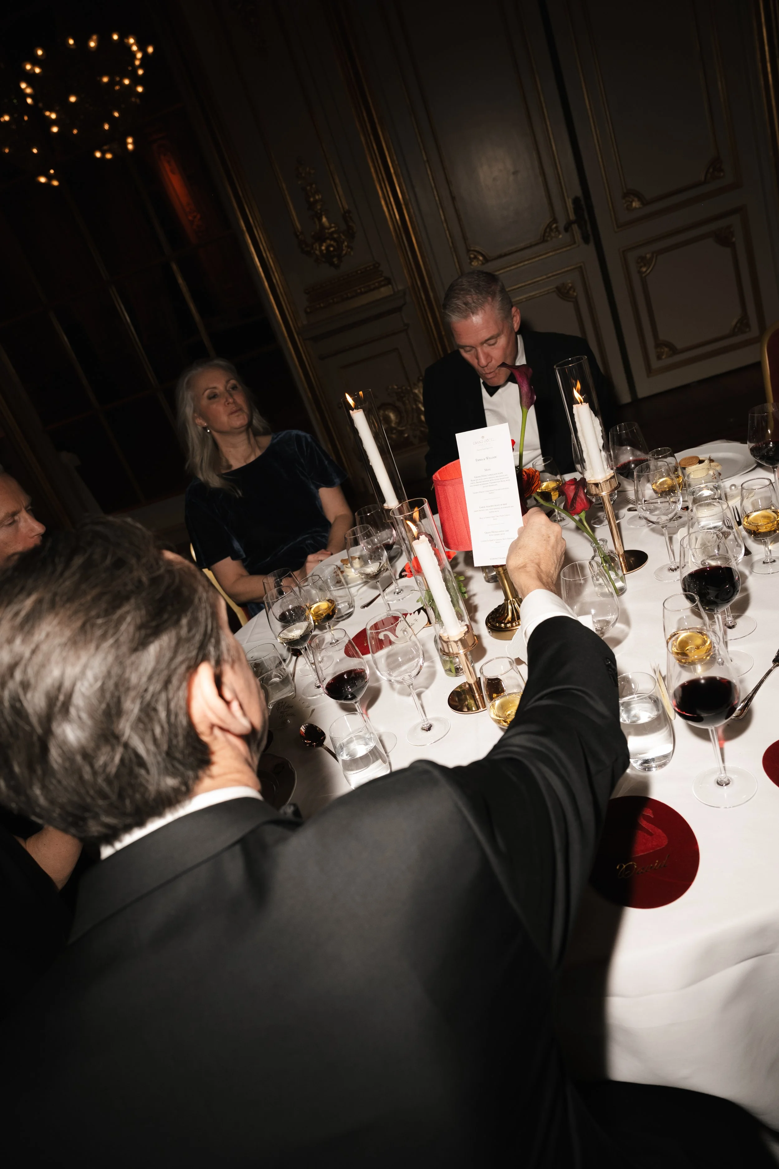 People dressed in formal attire having a toast at a candlelit dinner table in an elegant room with ornate decor and large candles.