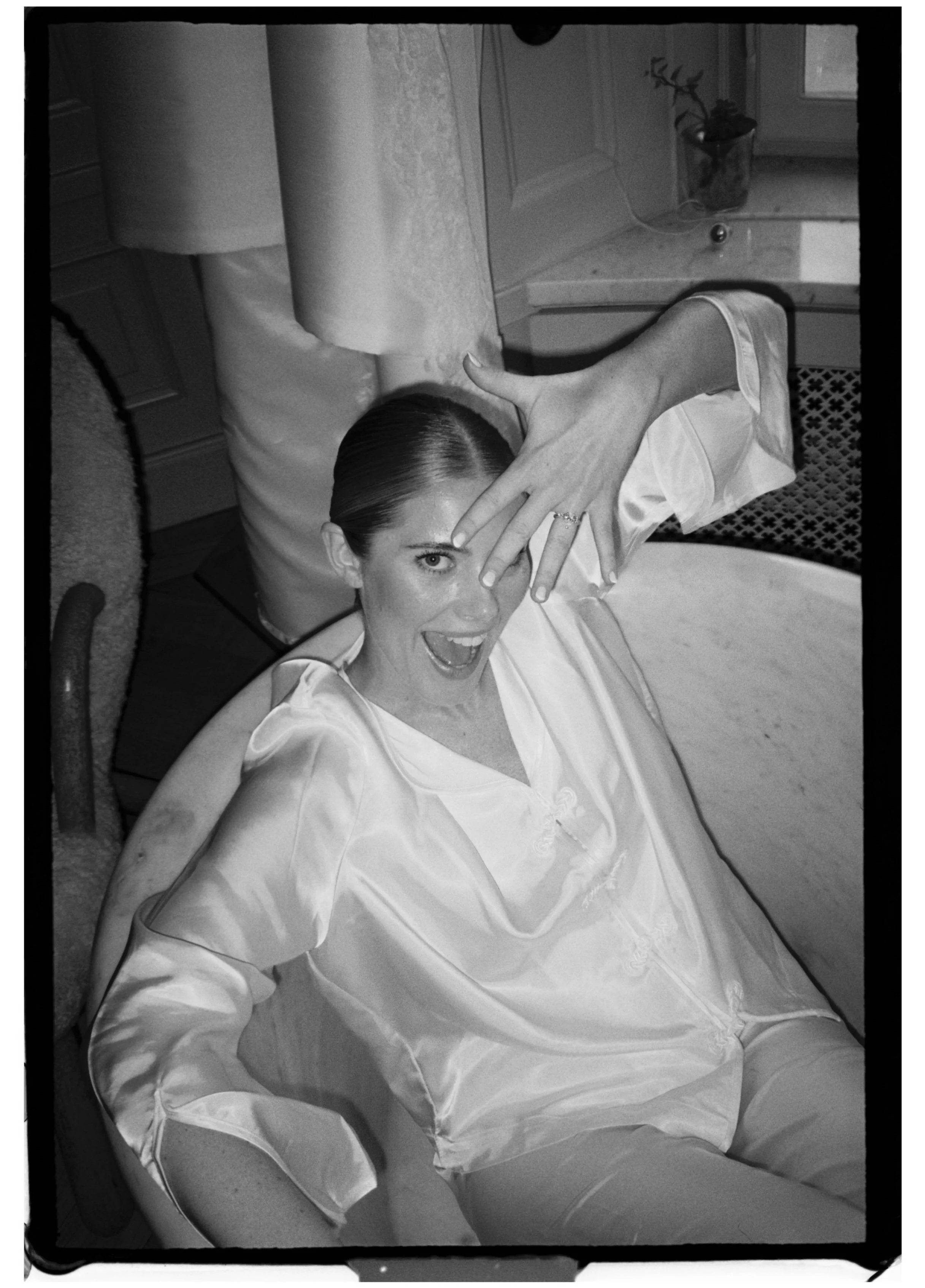 A woman lying in a bathtub, wearing a satin robe, playful and smiling, making a gesture with her hand over her face, in a bathroom with a window and decorative items.