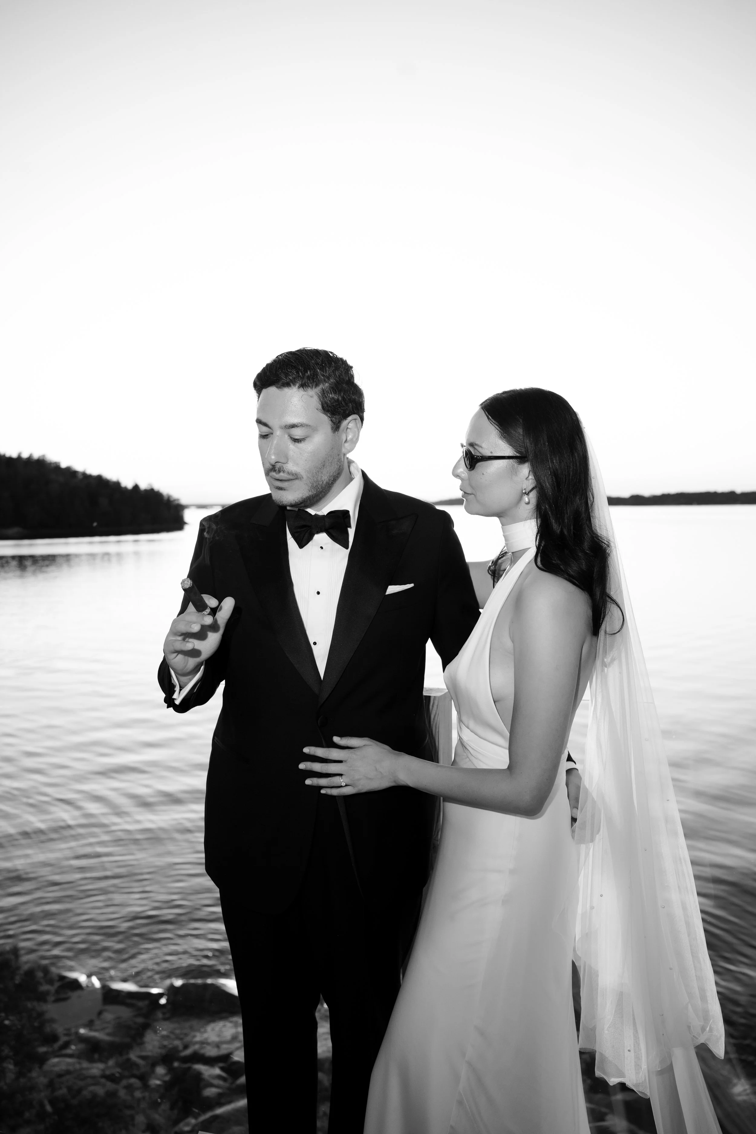 A black and white photo of a newlywed couple standing by a lake. The groom is in a tuxedo smoking a cigar, and the bride is in a wedding dress with glasses and a veil, looking at the groom.