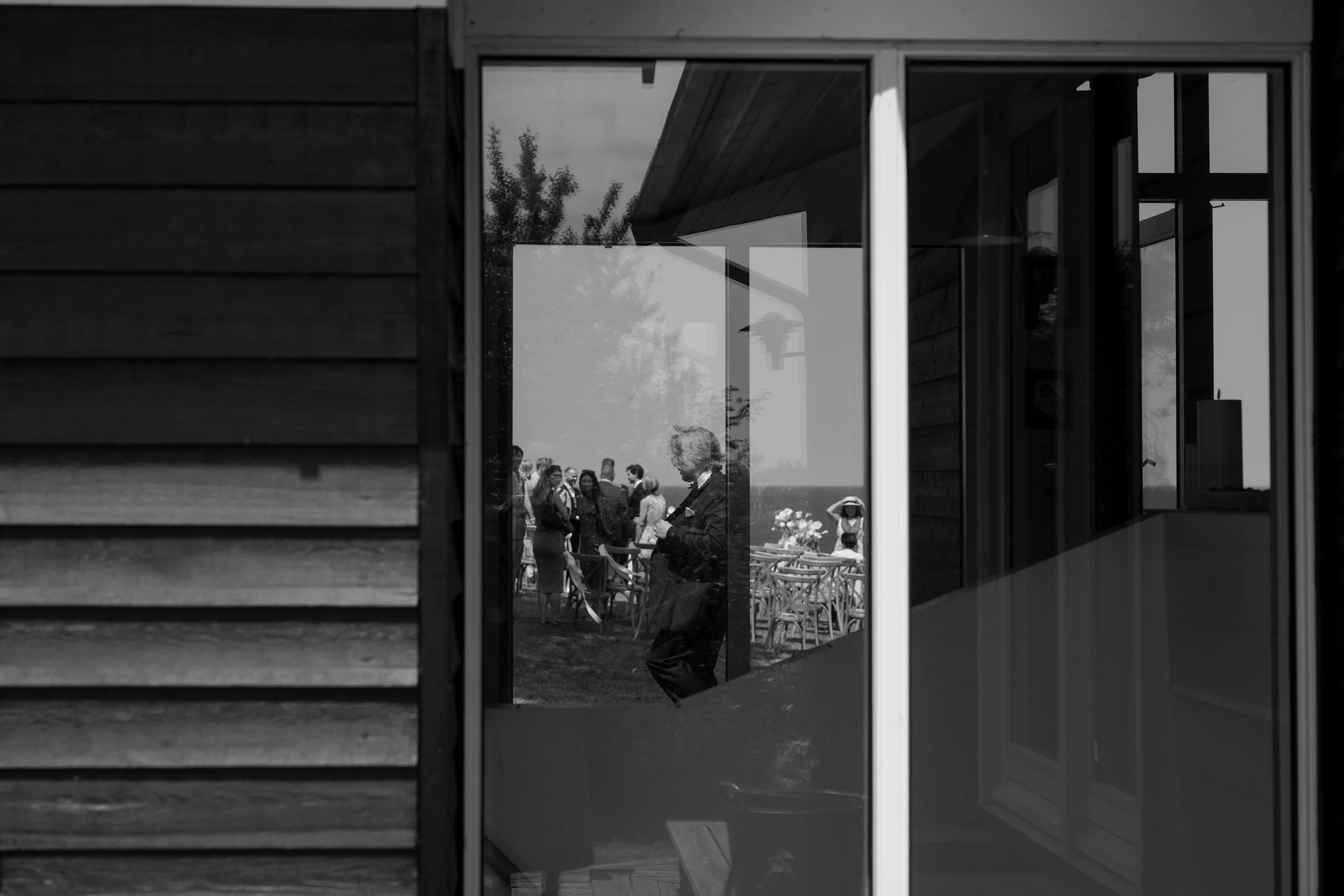 People gathered outdoors during a social event, viewed through a reflective glass window which shows part of the interior of a building.