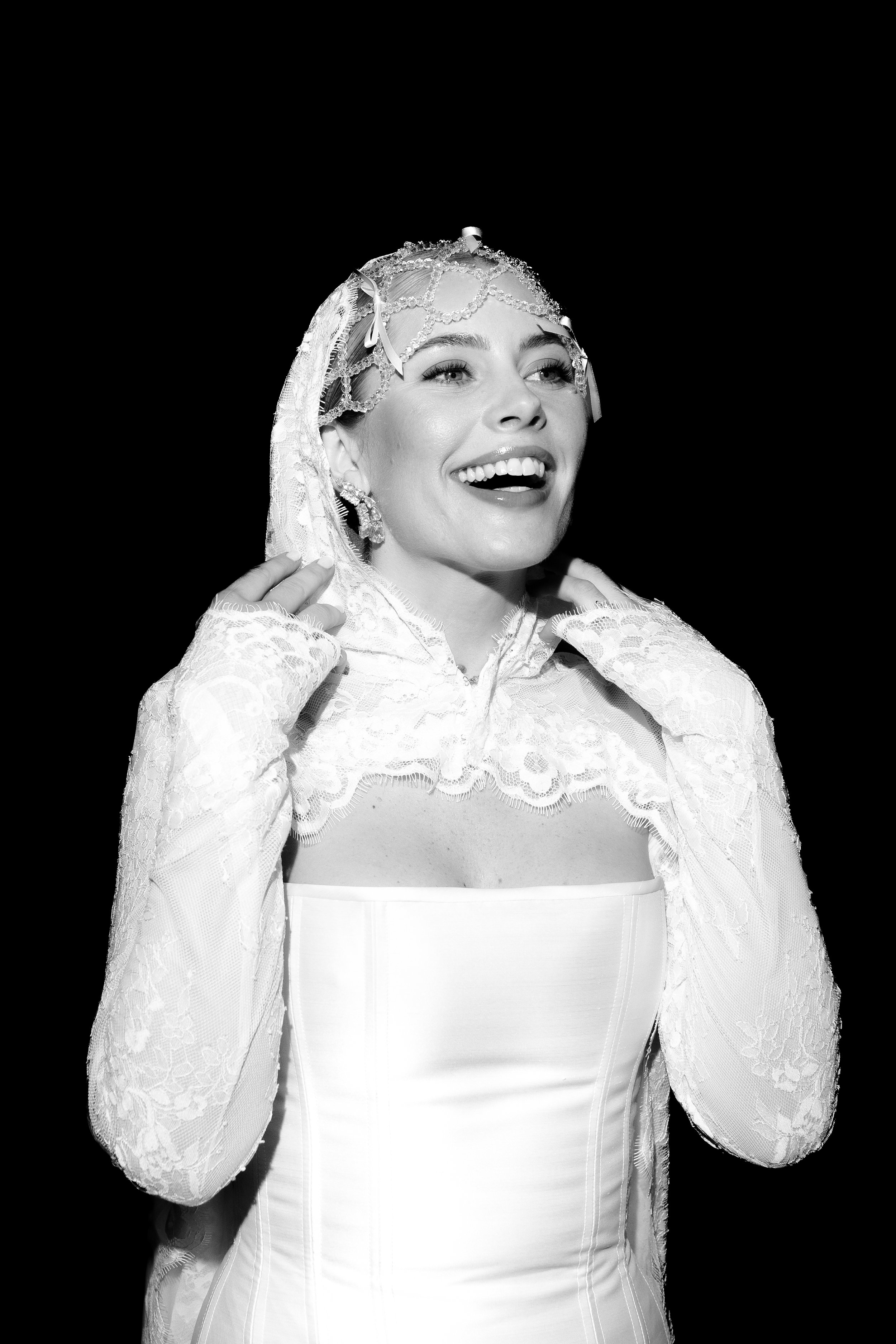 A woman in a vintage wedding dress and lace gloves, smiling with arms raised near her shoulders, wearing a lace veil and decorative headpiece, against a dark background.