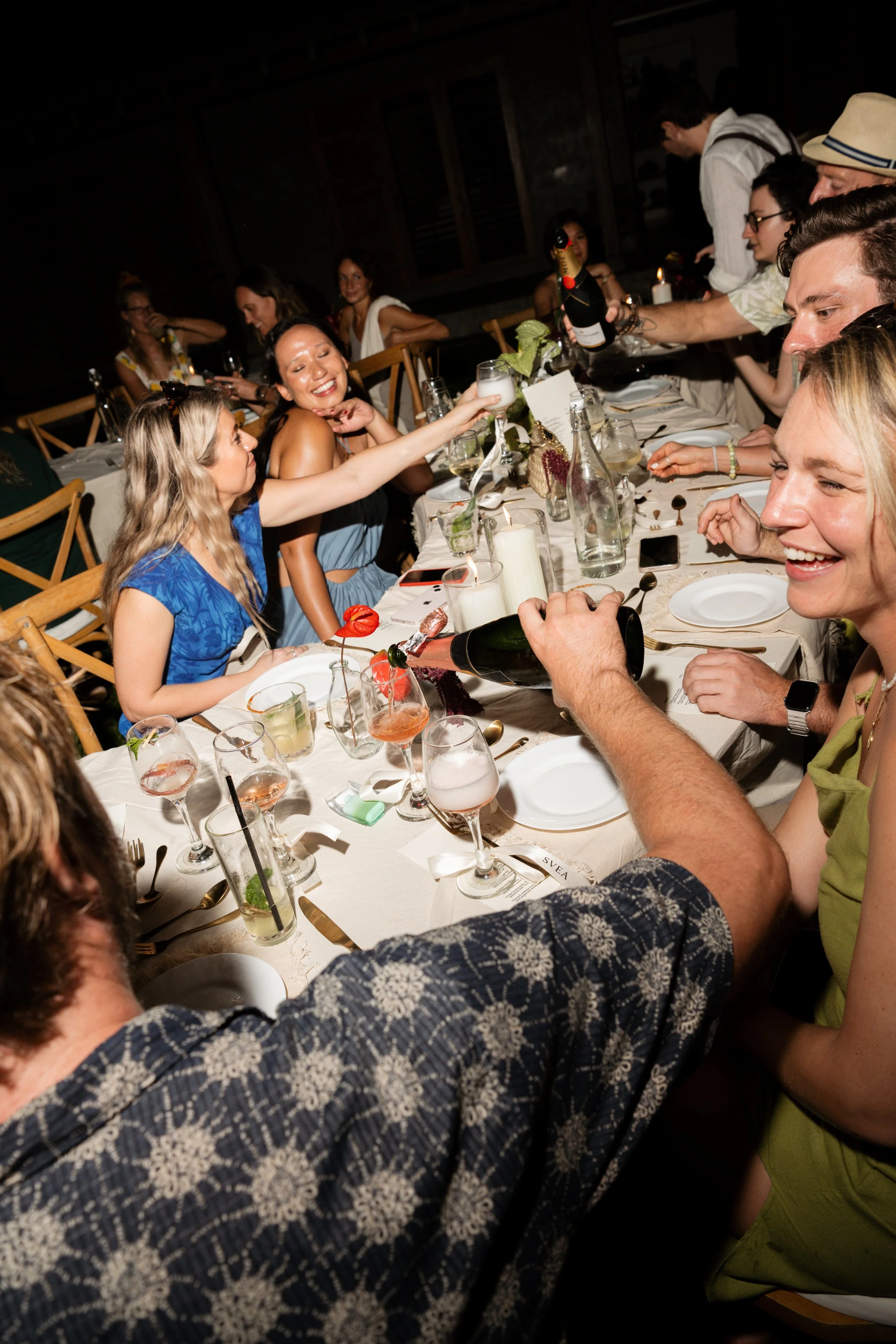 A group of people sitting around a dining table celebrating, with some guests pouring drinks and smiling, in a warmly lit setting.