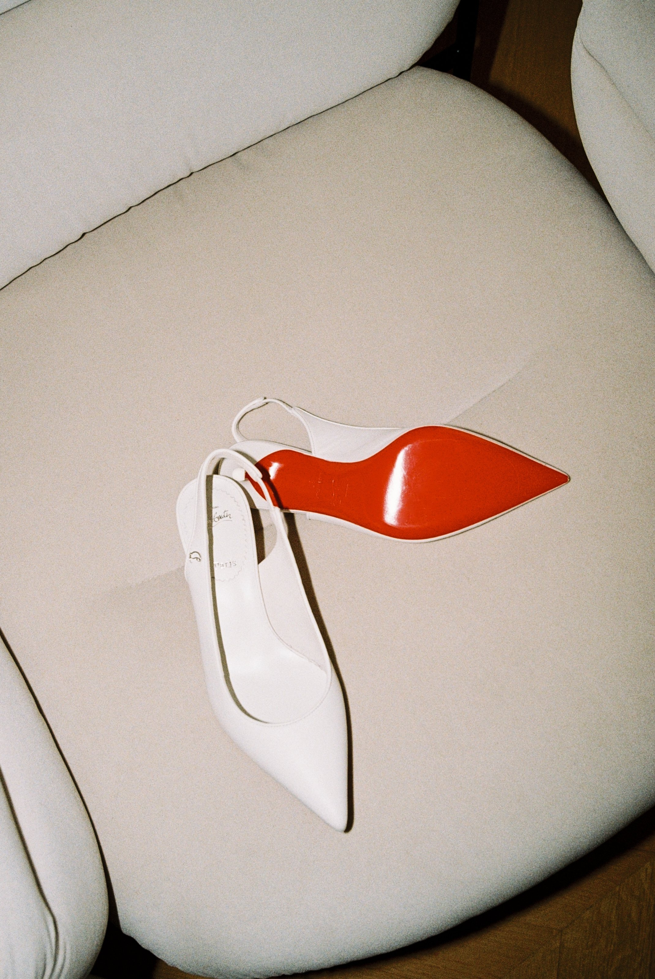 A white high-heeled shoe with a pointed toe and a strap, placed on a beige curved sofa. The shoe has a large red decorative element on the heel resembling a leaf or petal.