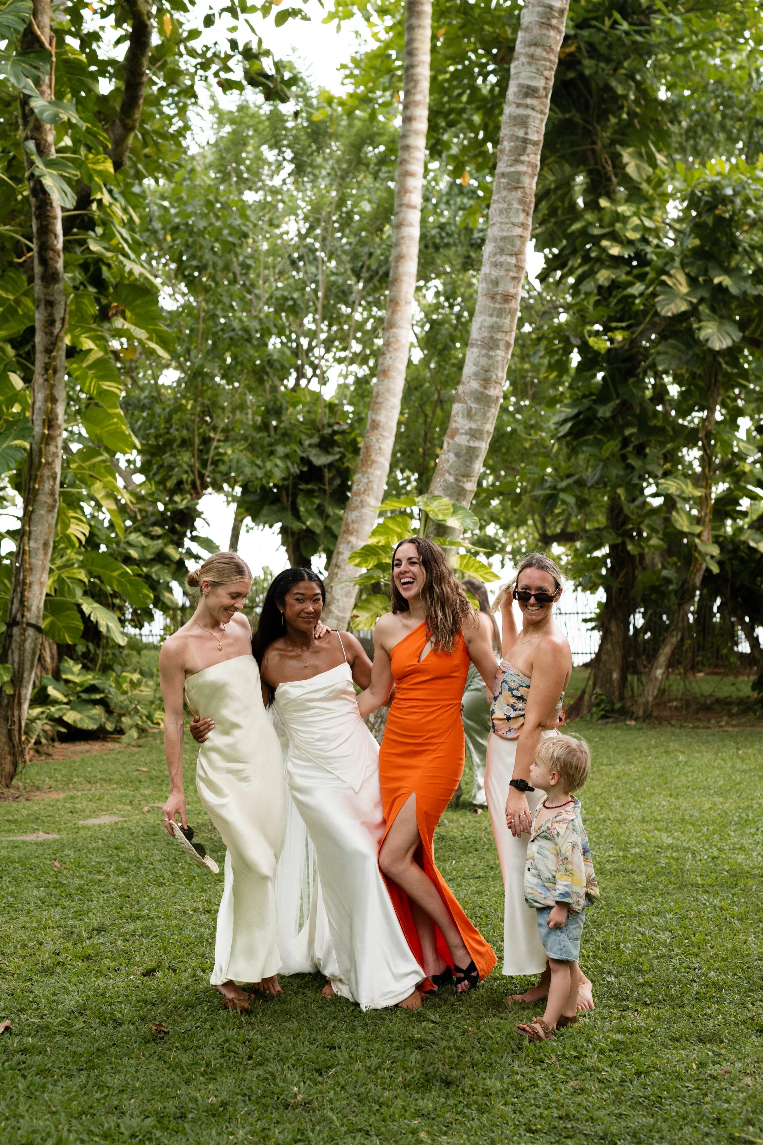 A group of five women and one little boy standing on grass in a lush green outdoor setting, smiling and enjoying themselves.