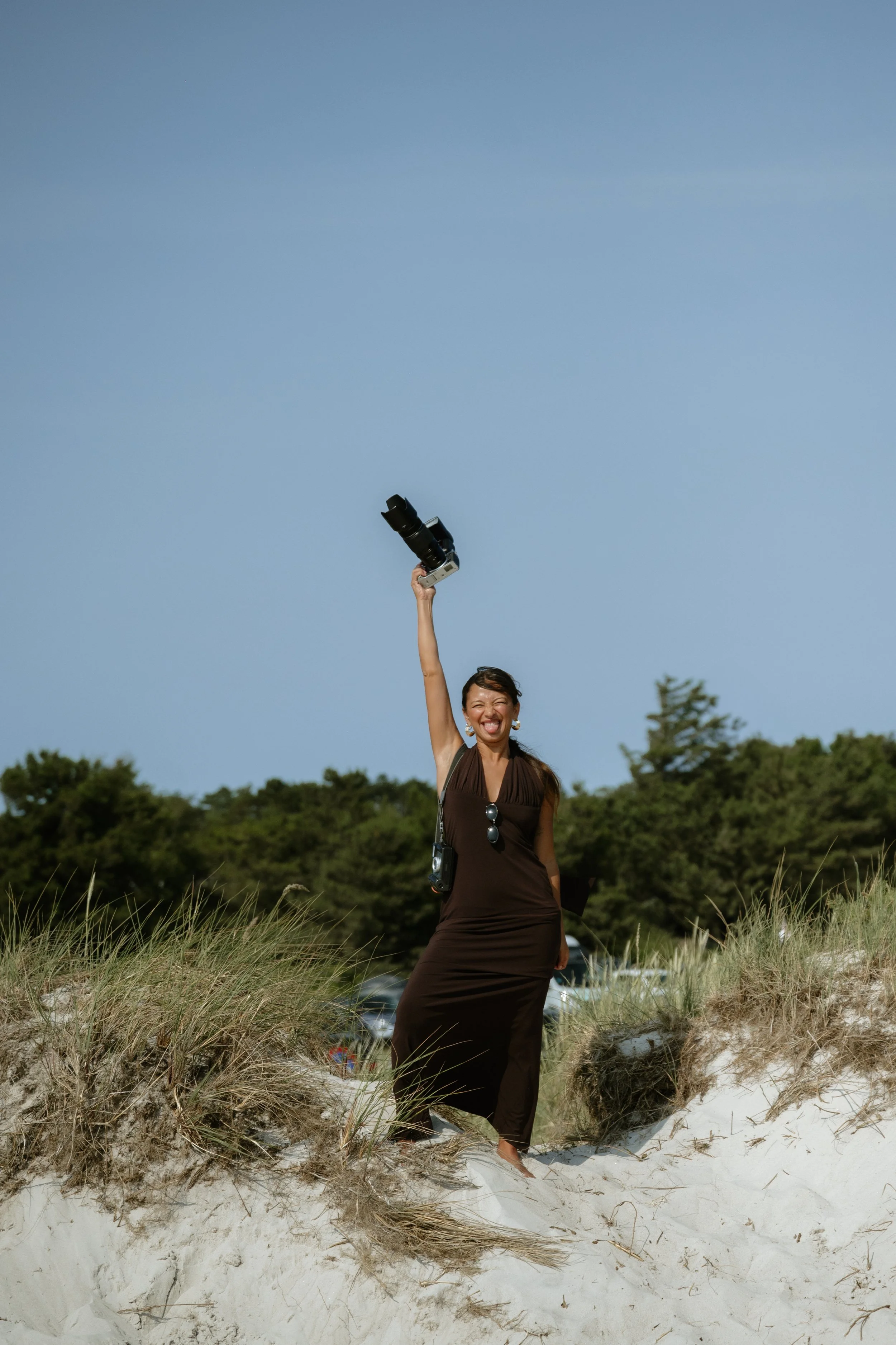 A woman in a brown dress standing barefoot on a sandy dune at the beach, holding a camera with a zoom lens high in the air, smiling joyfully, with green trees and a clear blue sky in the background.