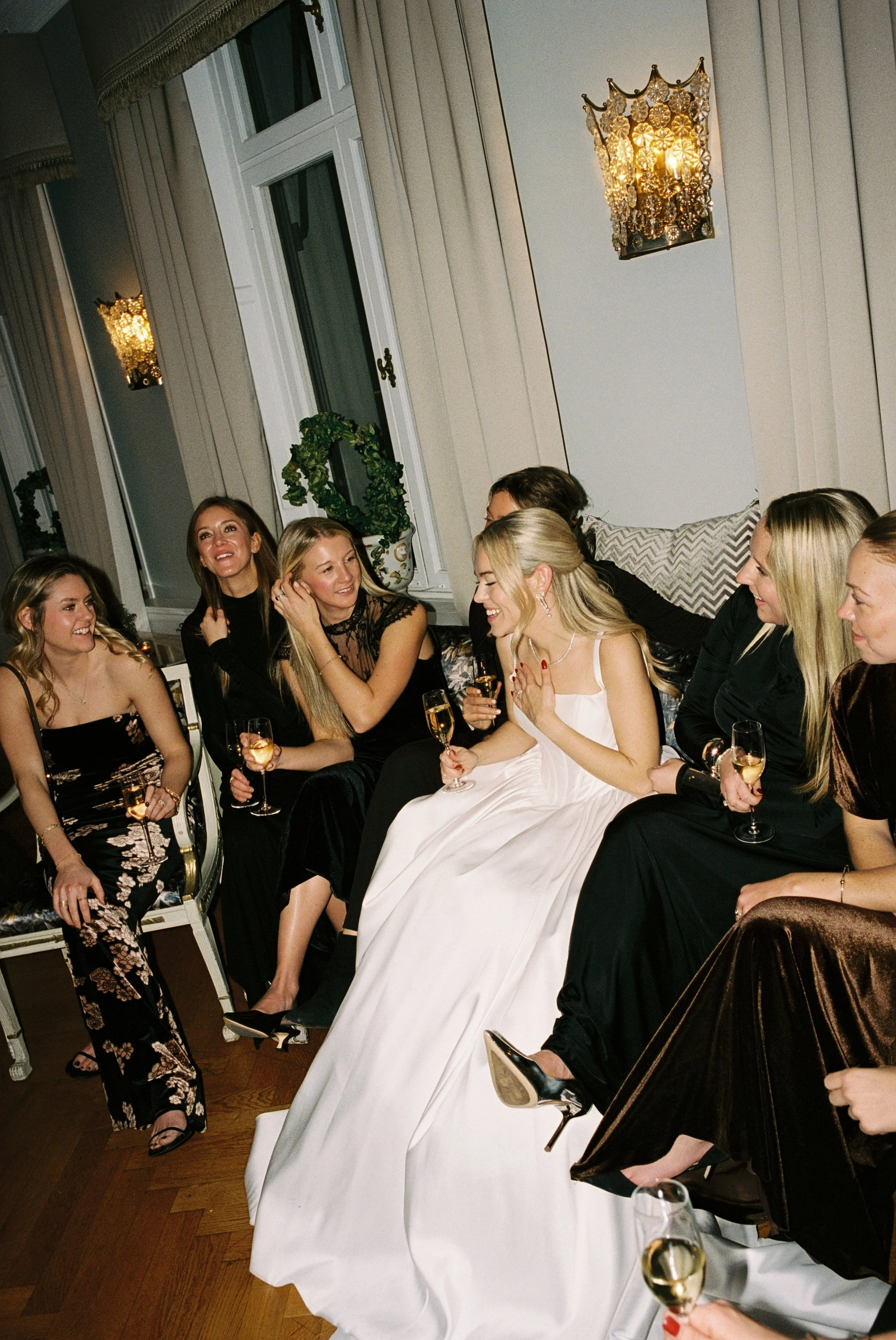 Women in an elegant living room celebrating, with some women wearing black dresses and one woman in a white gown, holding champagne glasses and smiling.