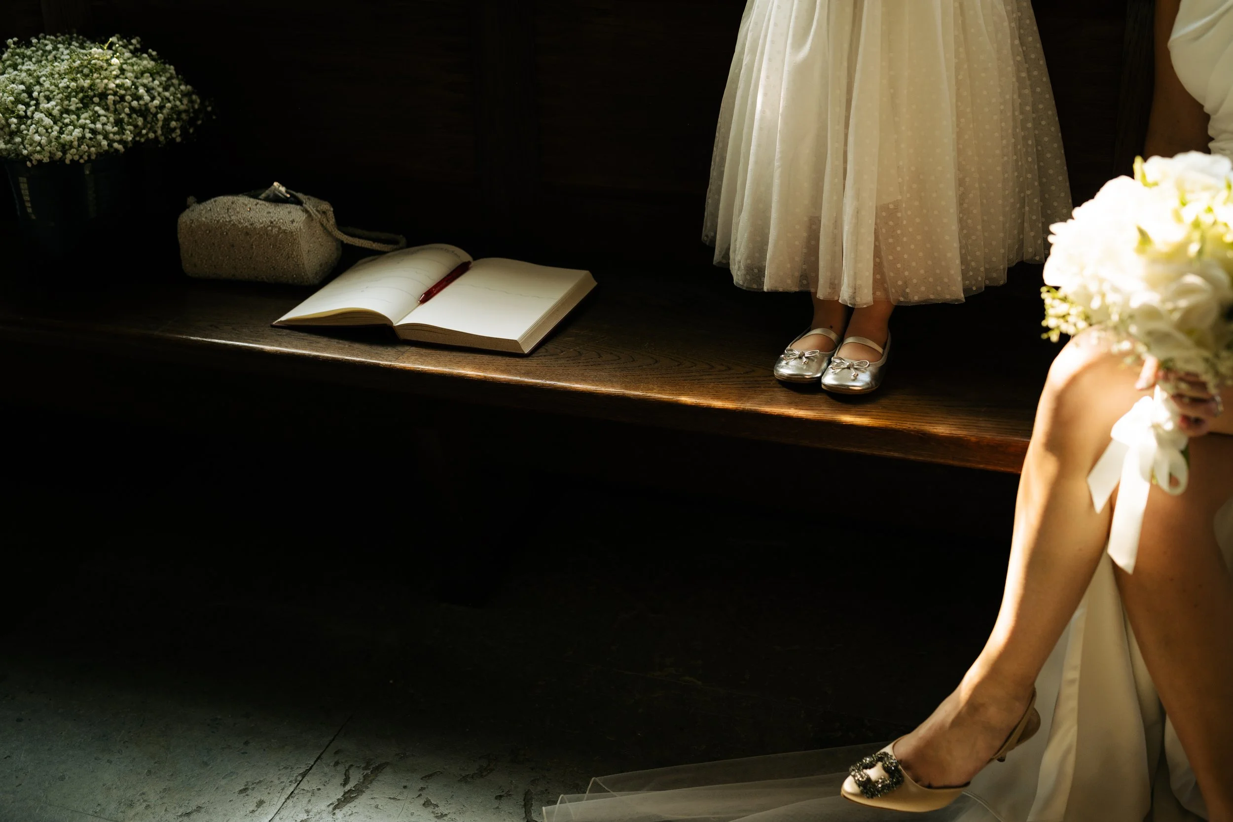 A wedding scene showing a bride seated with her leg crossed, holding a bouquet of white flowers, and a young girl standing on a wooden bench wearing a cream-colored dress and silver shoes. An open guestbook and a purse rest on the bench.