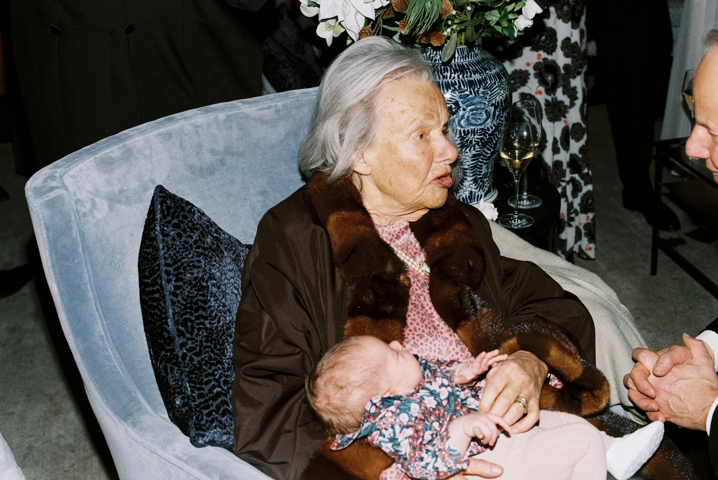 An elderly woman sitting on a light blue armchair with a black patterned cushion, holding a small child on her lap. The woman has gray hair, is dressed in a brown jacket with a fur collar, and a pink patterned blouse. There is a large floral arrangem