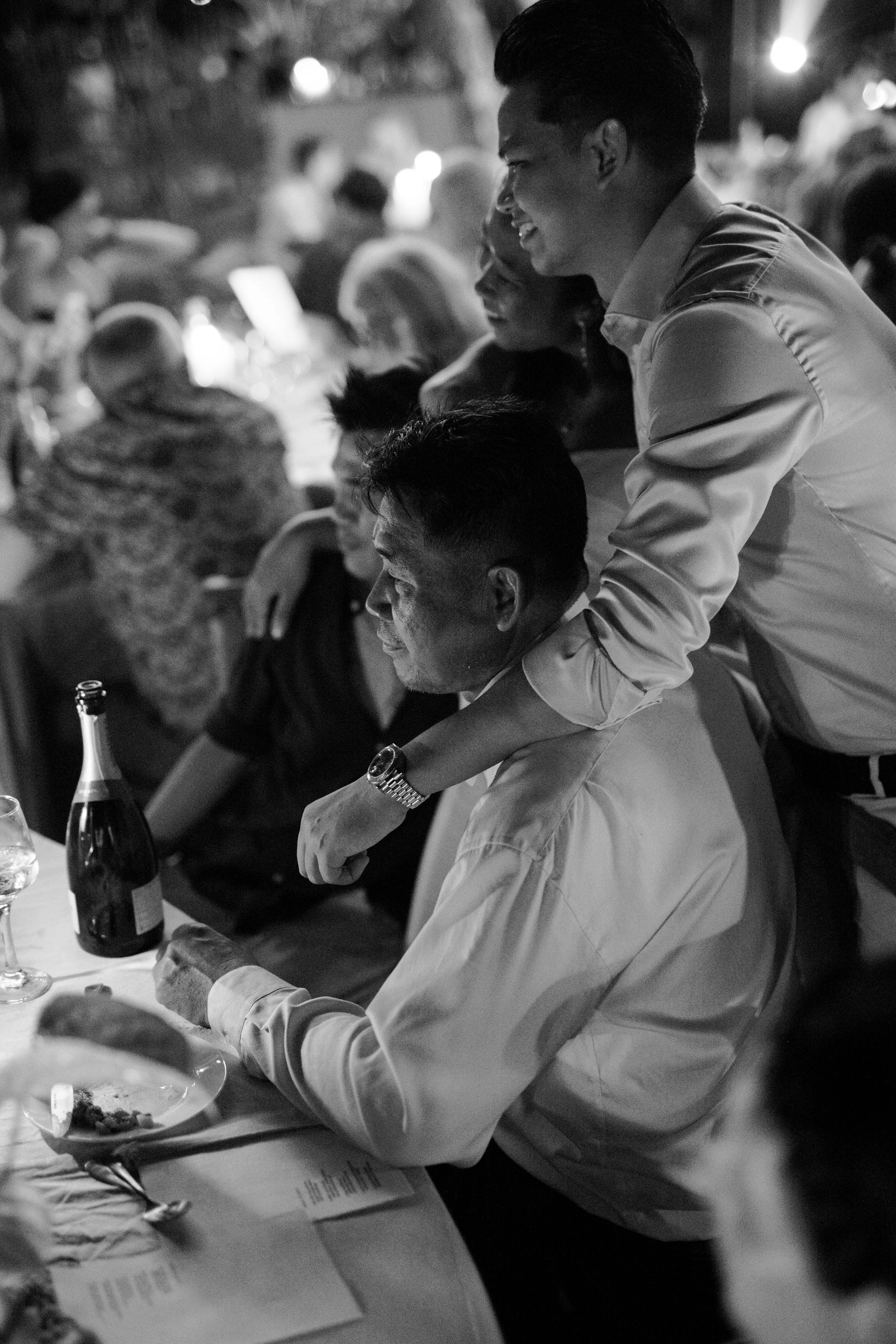 People enjoying a celebration dinner, with one man whispering into another's ear at a restaurant or party setting.