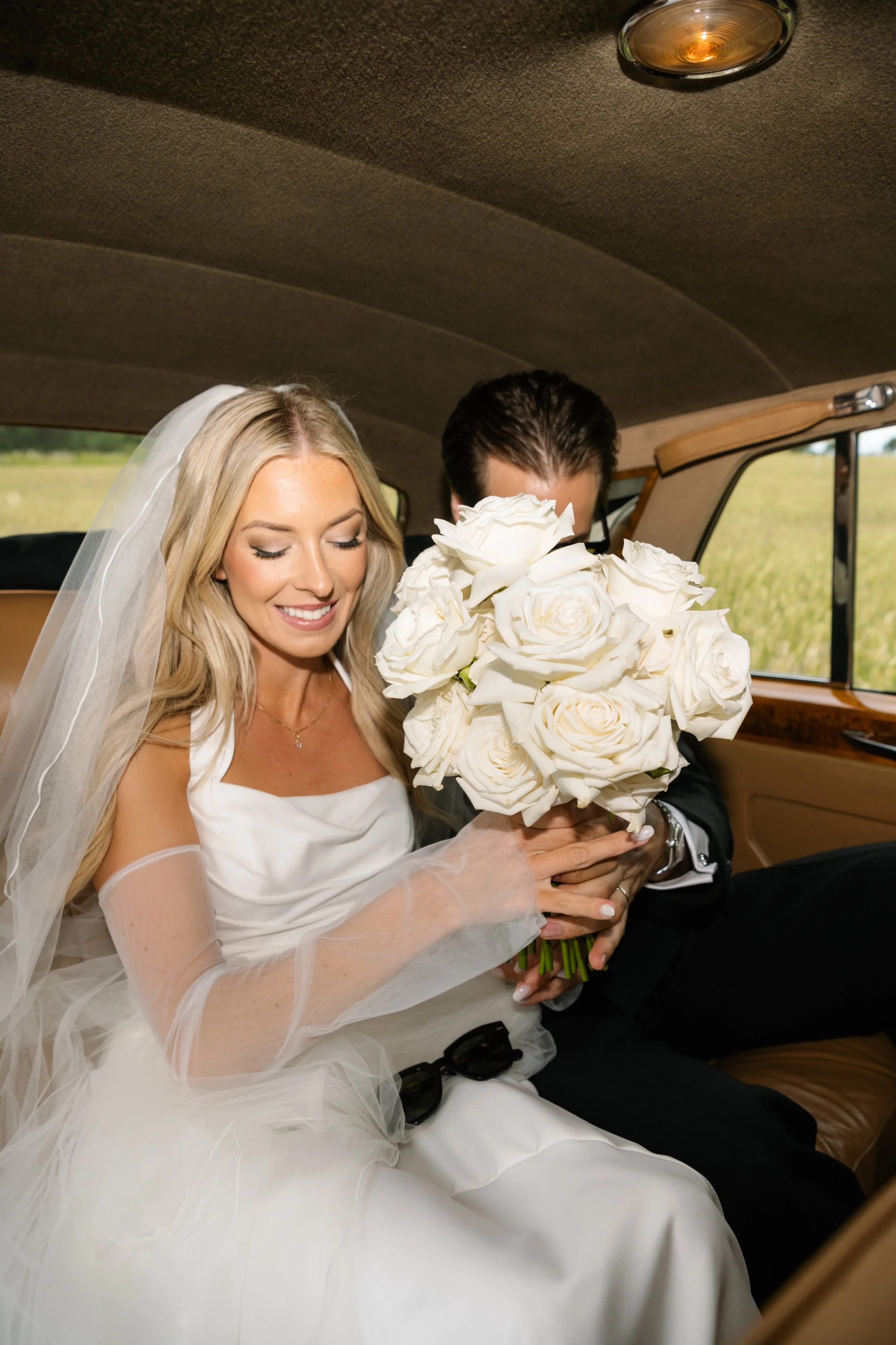 Bride in wedding dress holding a bouquet of white roses, sitting inside a vehicle with a man partially visible behind the bouquet.