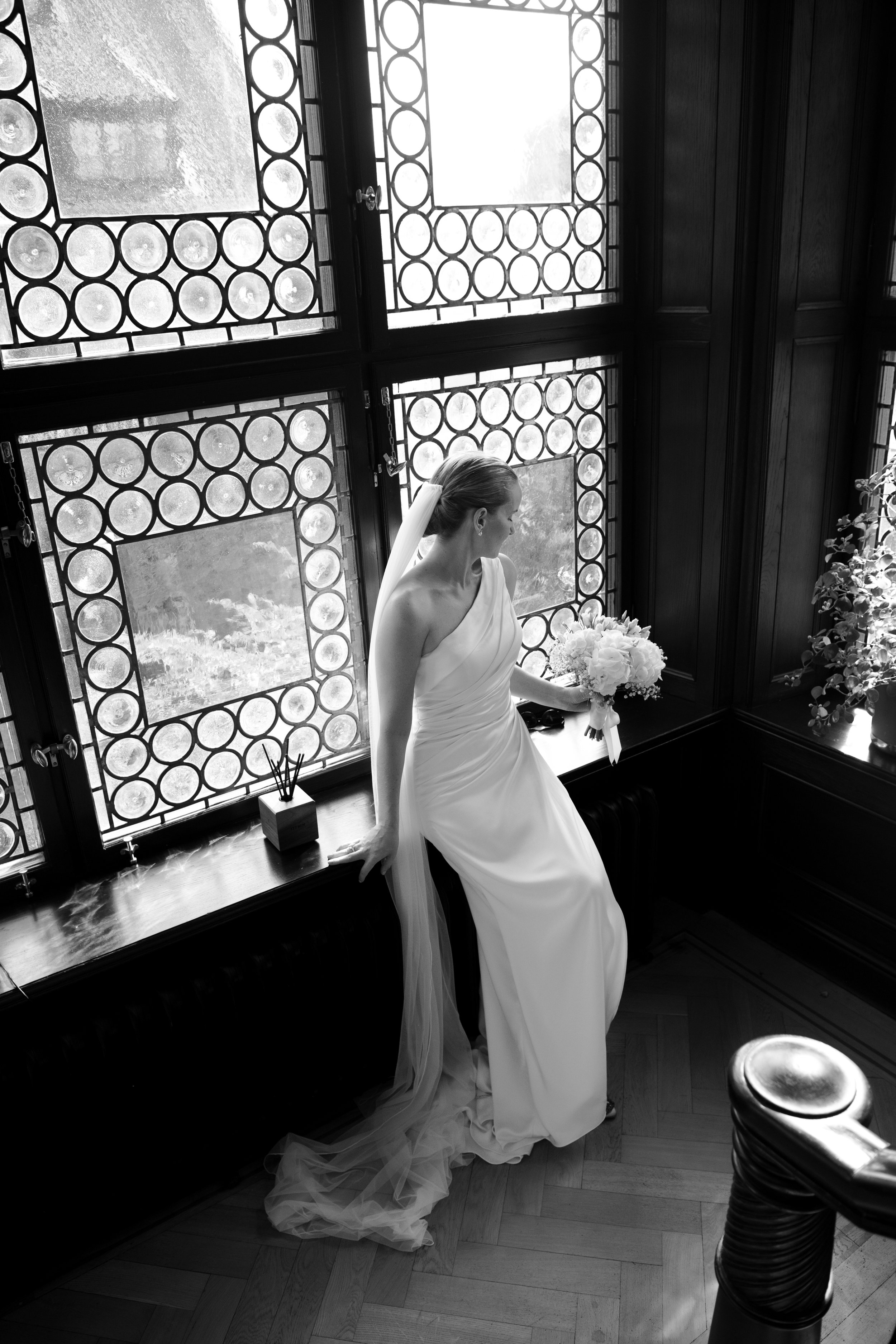A bride in a white wedding gown holding a bouquet, sitting on a windowsill in a room with stained glass windows and dark wooden paneling.