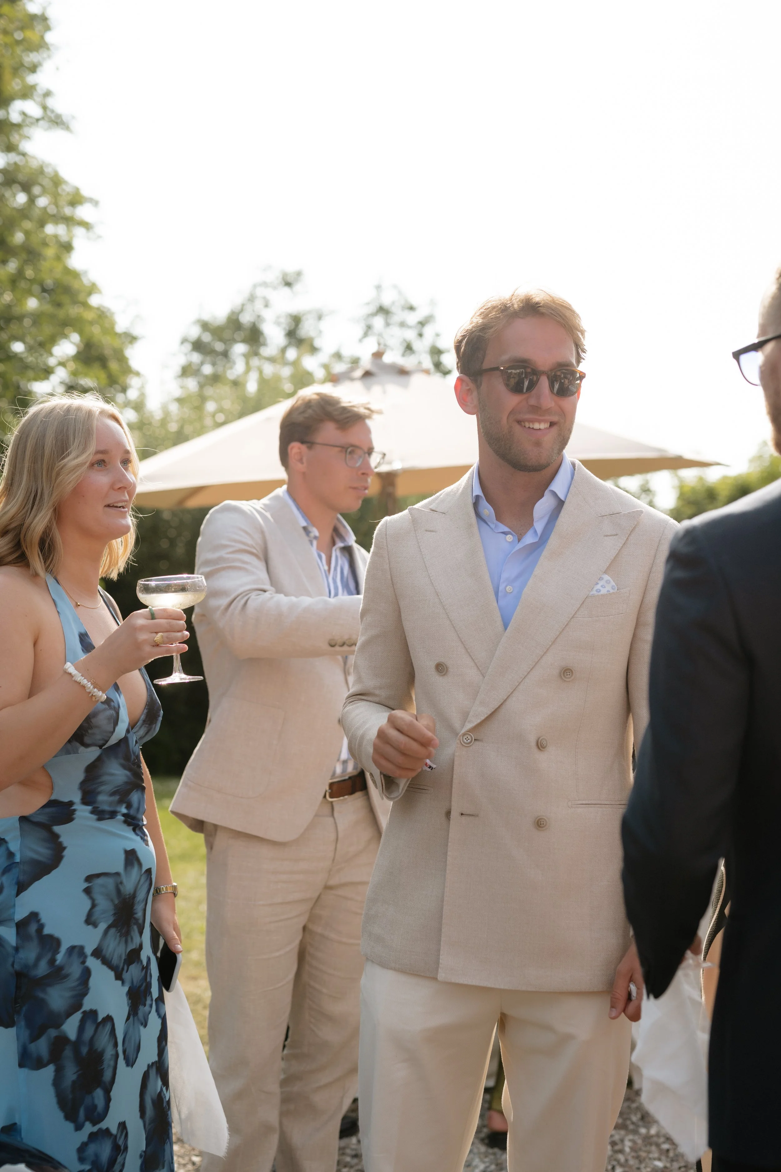 People dressed in formal and semi-formal attire socializing outdoors at a gathering with a large umbrella in background