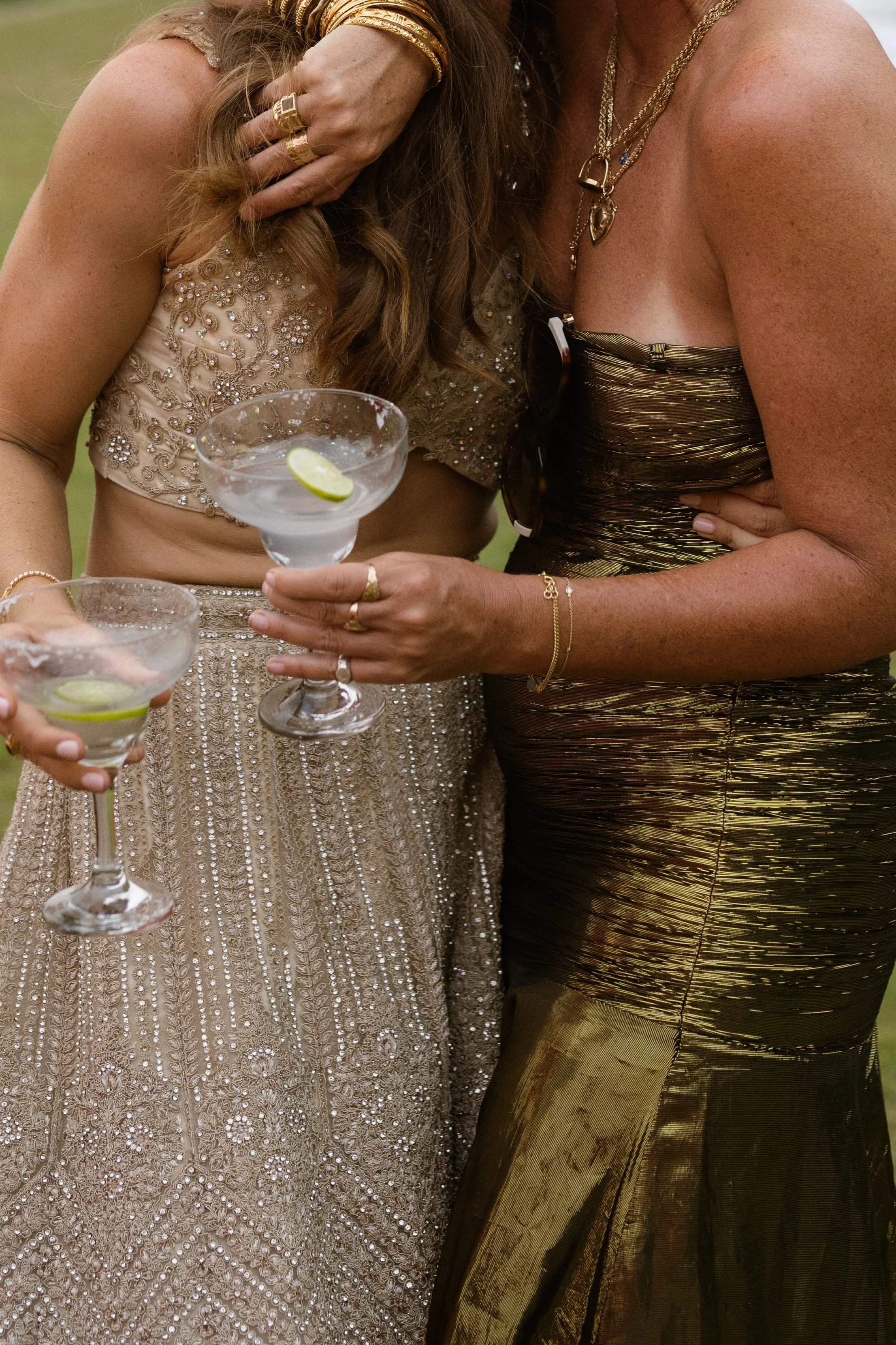Two women at a celebration holding cocktails with lime slices, dressed in elegant, shimmering dresses, wearing jewelry.