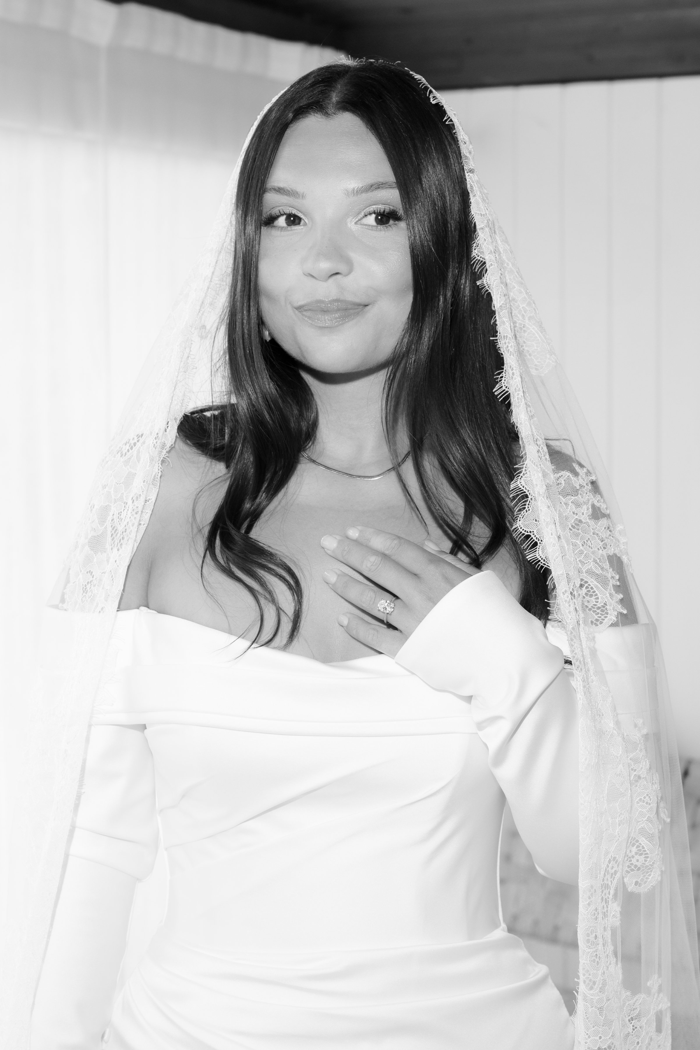 A woman in a wedding dress with a veil, touching her chest with her left hand, looking slightly to her right.