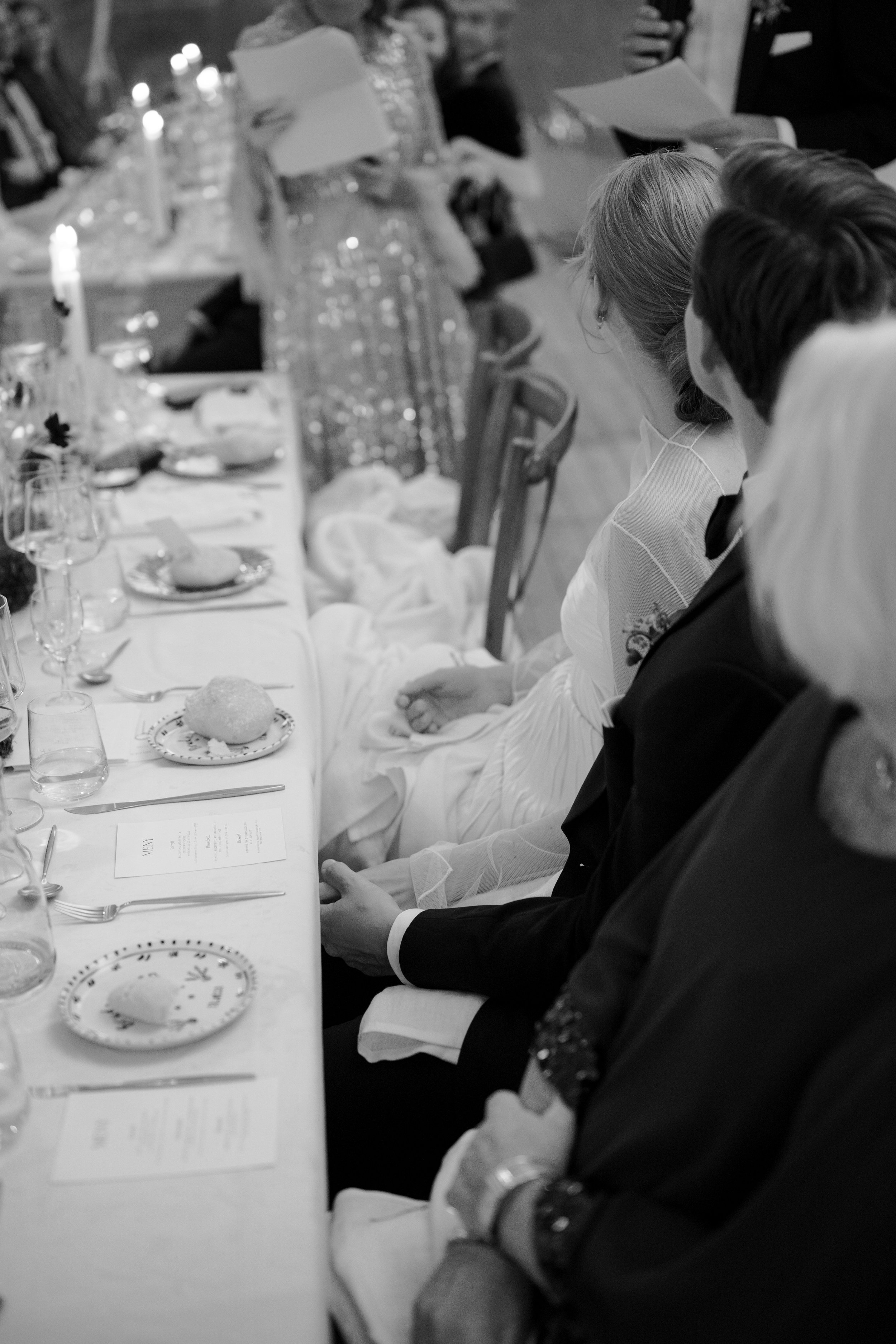 Black and white photo of a wedding reception table with guests seated, including a bride in a white dress and a groom in a tuxedo, viewed from behind.