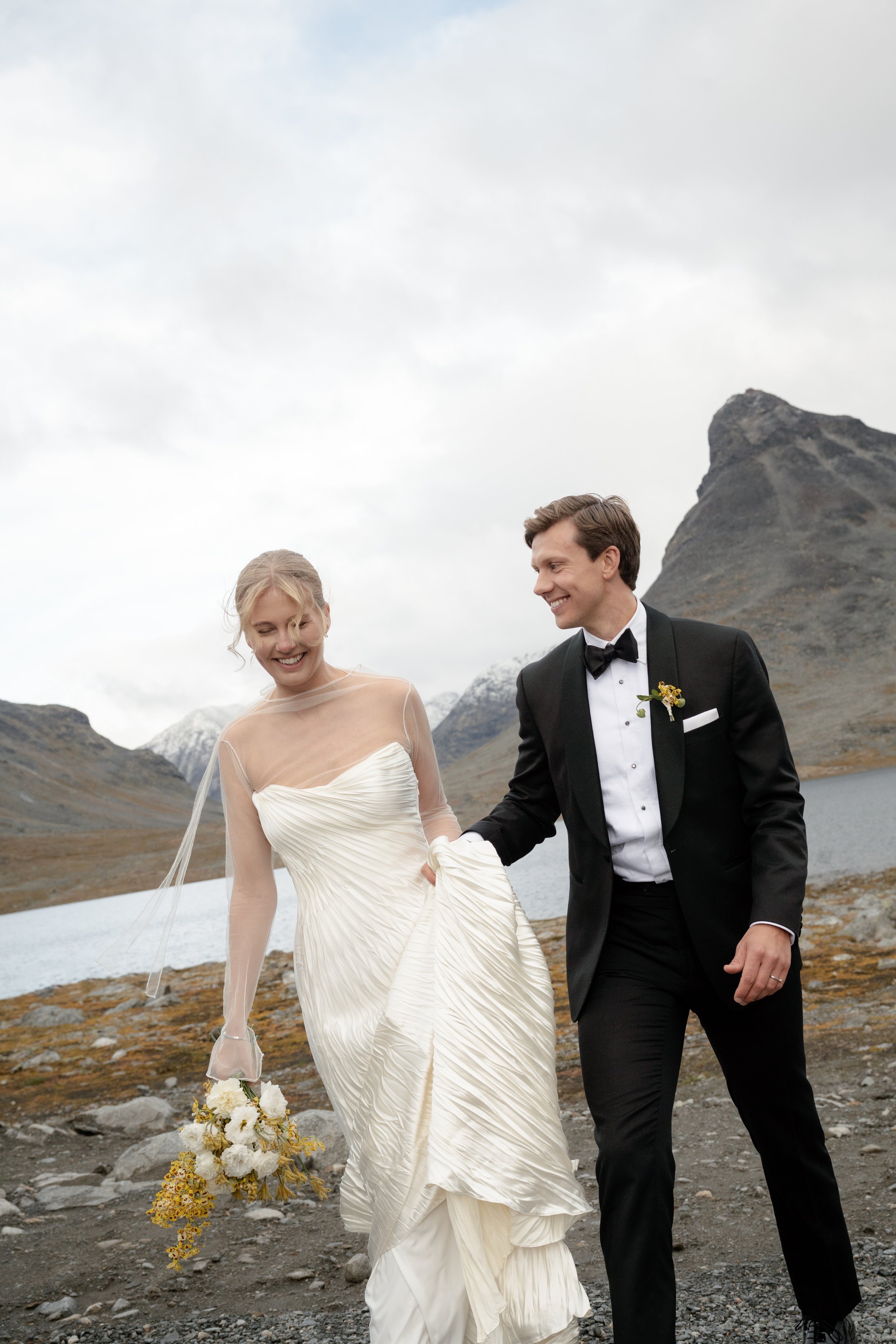 A bride and groom walking outdoors near a lake, with mountains in the background, dressed in wedding attire, smiling and holding hands.