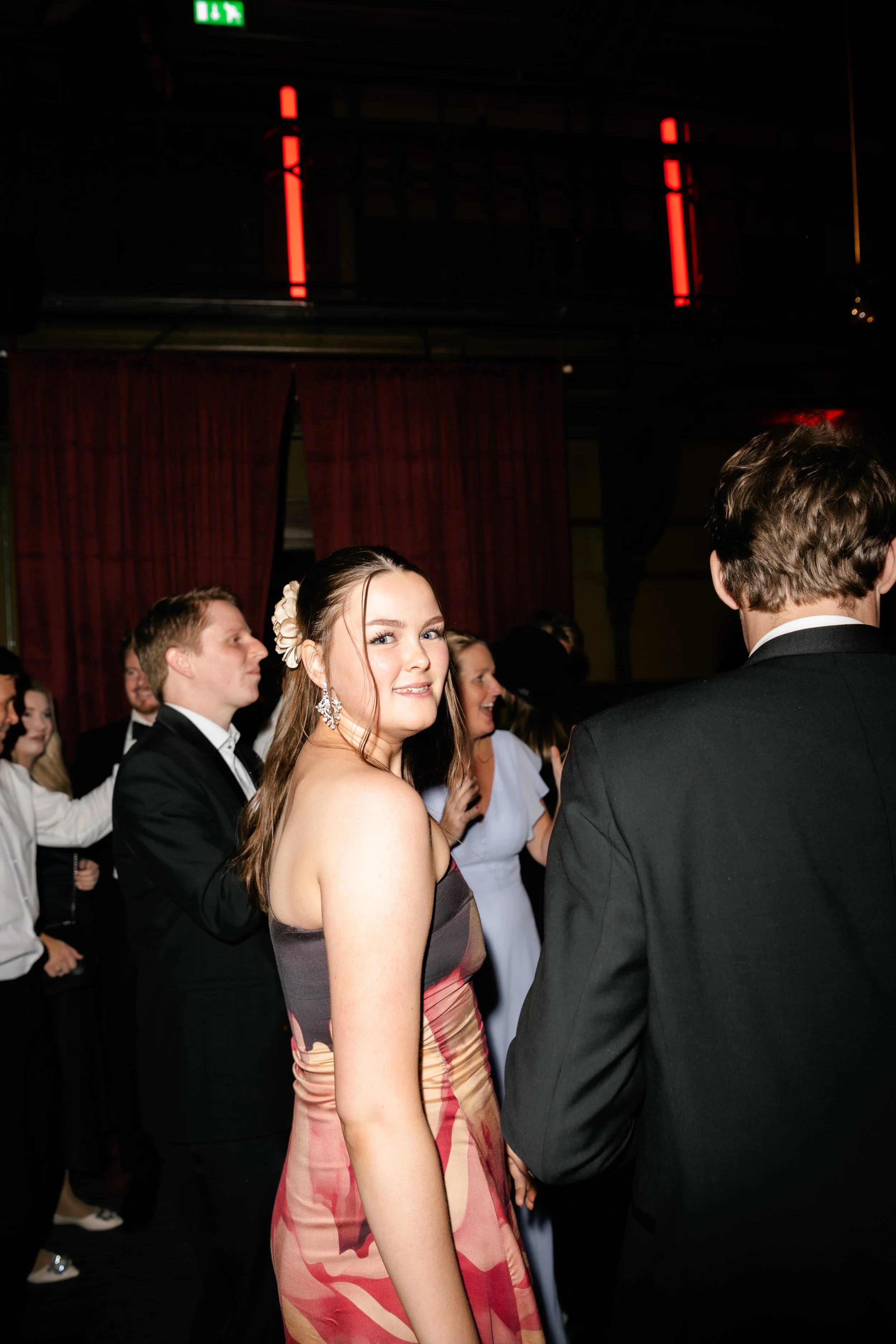 A woman in a strapless dress smiling at a party or formal event, with several other people dancing or socializing in the background, indoors with dark lighting and red curtains.
