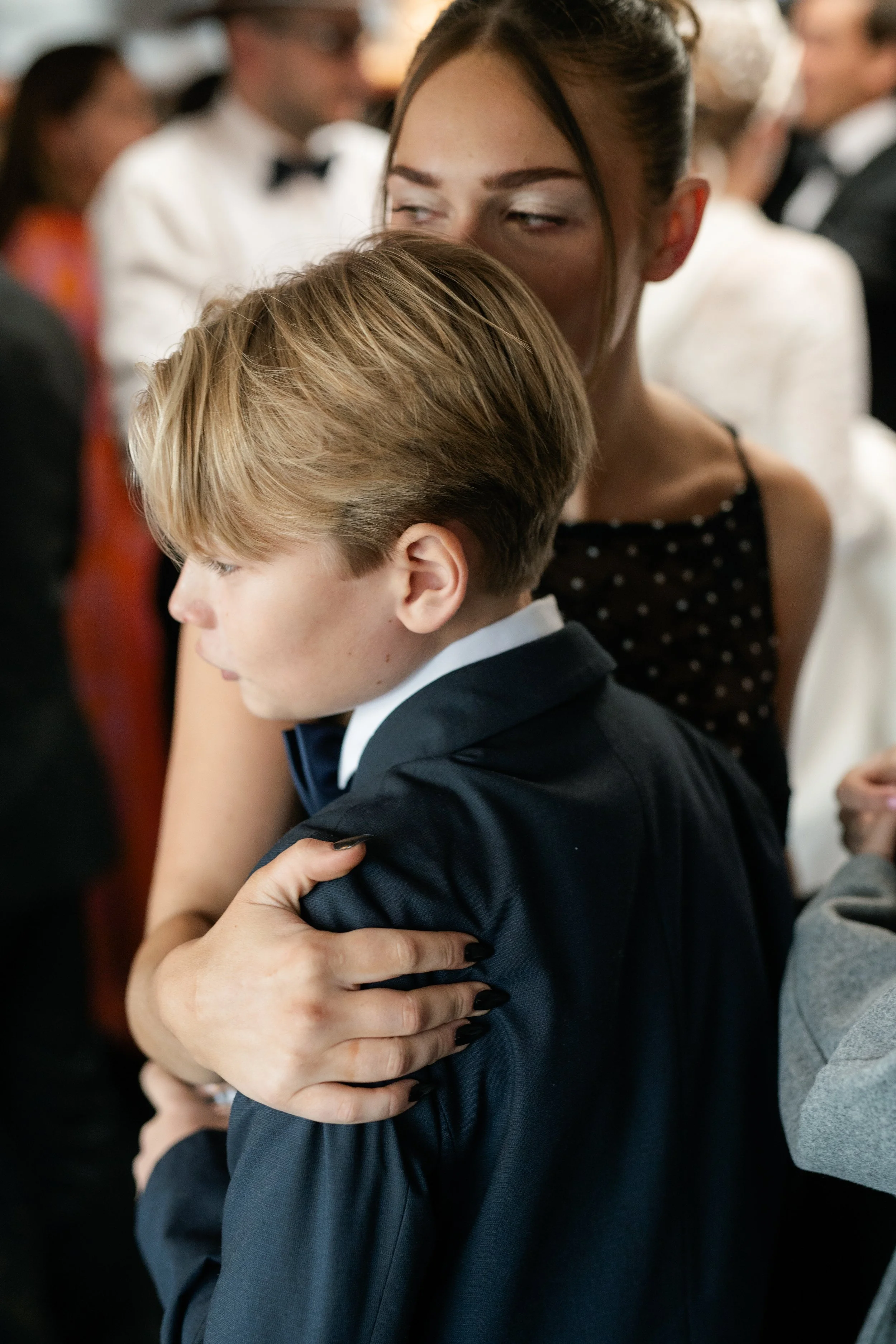 A woman and a young boy are hugging at a formal event, with people in formal attire in the background.