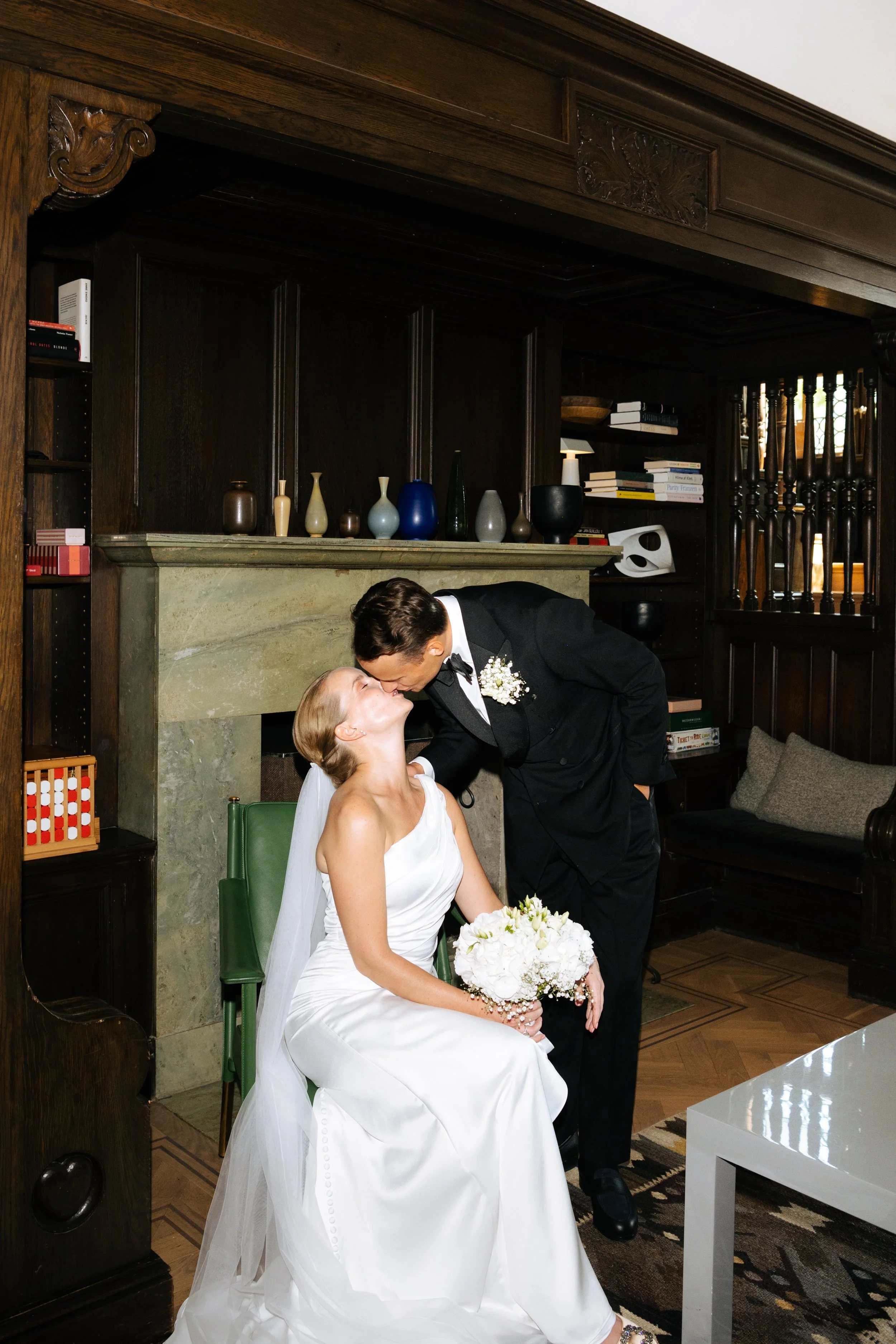 A bride and groom share a kiss in front of a fireplace in a cozy, elegant room.