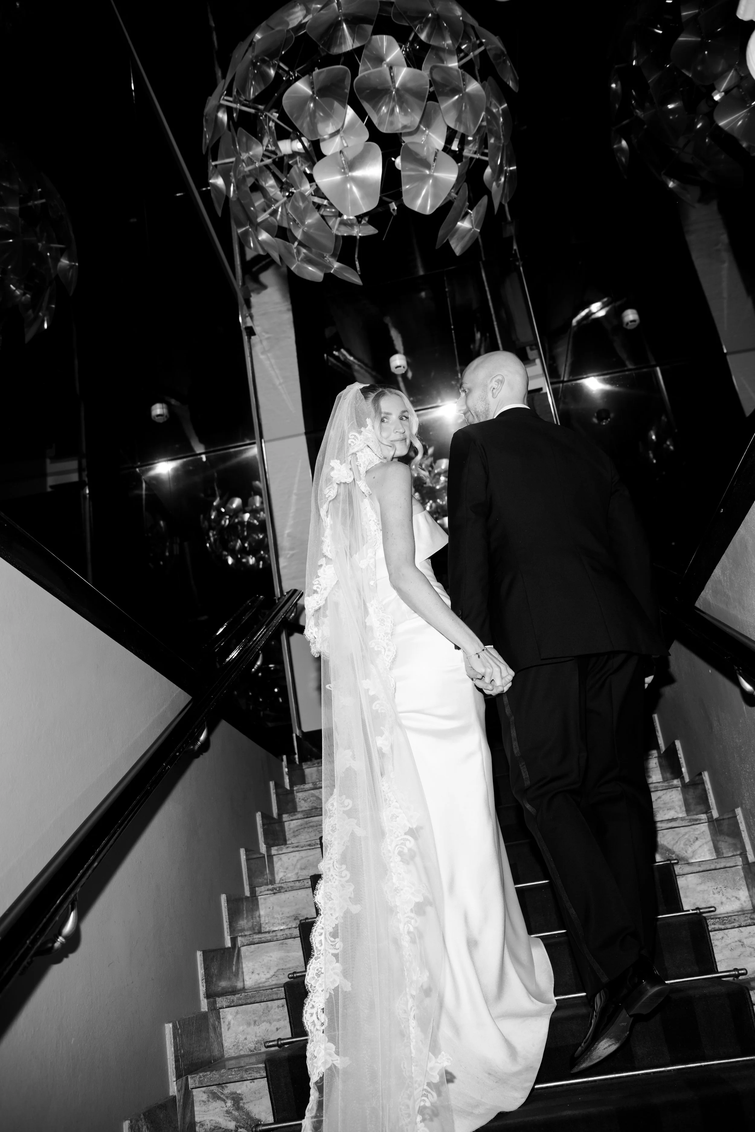 Bride and groom holding hands on a staircase, with the bride wearing a veil and wedding gown, and the groom in a black suit, inside a modern venue with ceiling chandeliers and reflective walls.