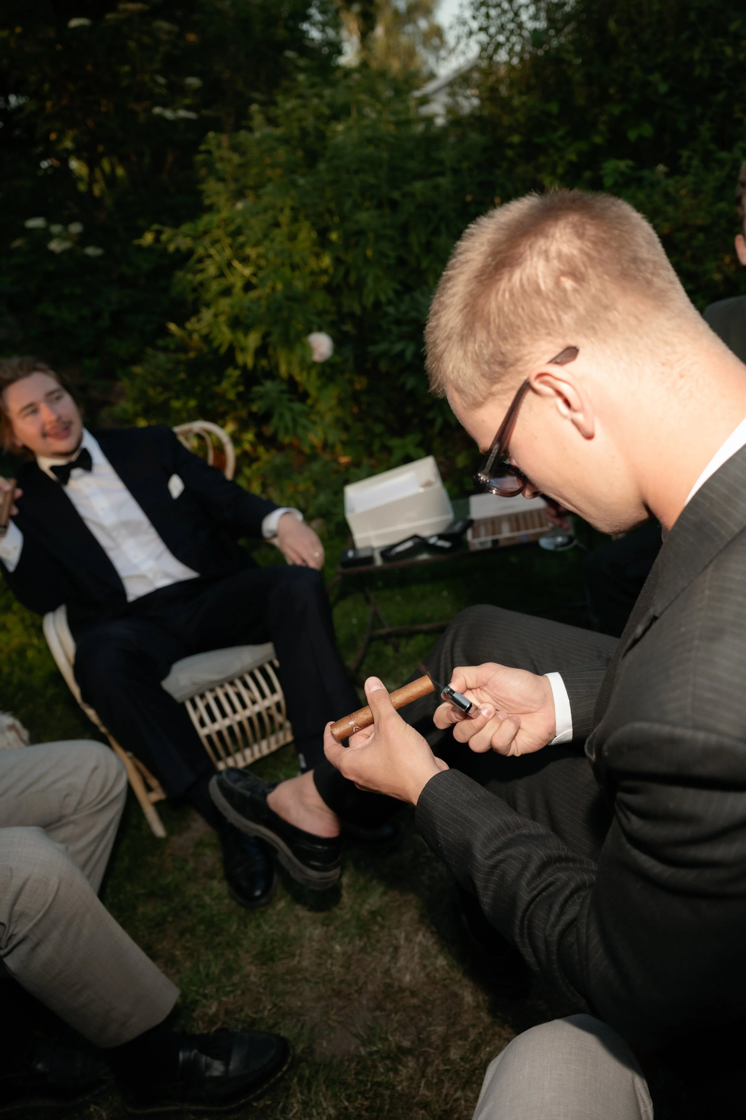 A man in a black suit and glasses is sitting outdoors, holding a cigar and a lighter. In the background, a man in a tuxedo is sitting on a chair, smiling and gesturing, with greenery and outdoor furniture around.