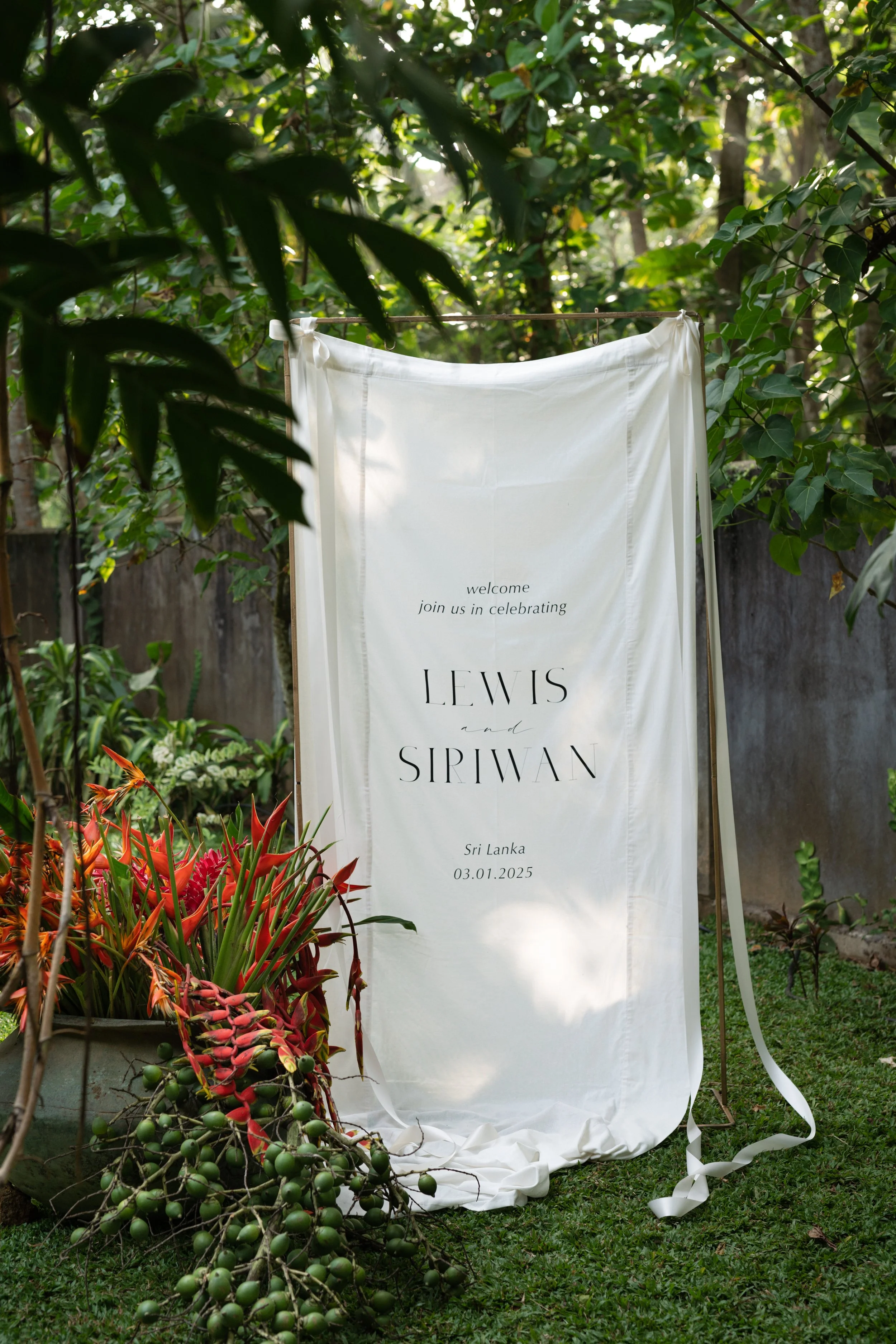 Wedding welcome sign with black text on white fabric, standing outdoors among green foliage and colorful plants, displaying the names Lewis and Sipwan, the location Sri Lanka, and the date 03.01.2025.