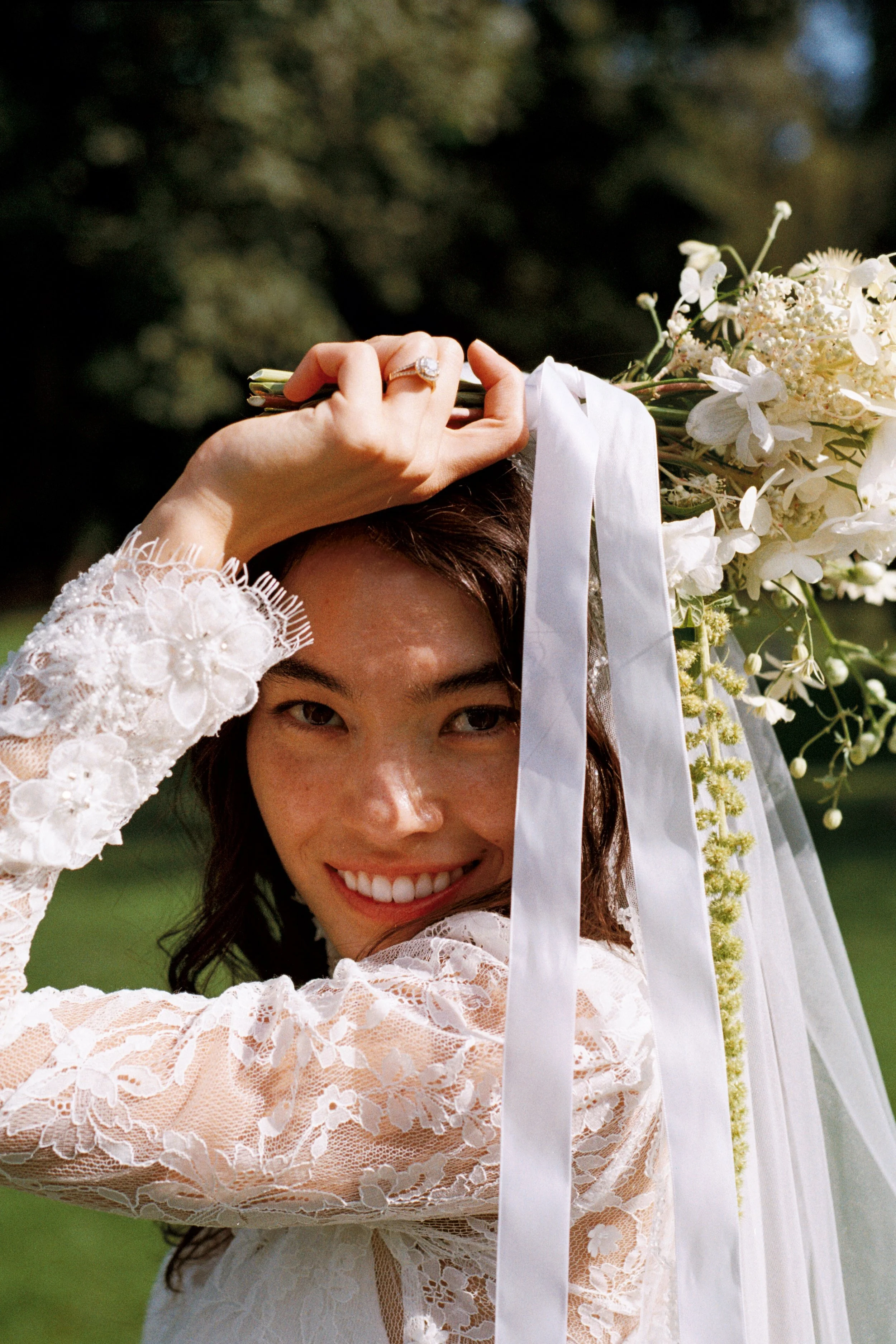 A woman in a lace wedding dress holding a bouquet of white flowers and ribbons, smiling outdoors with trees and greenery in the background.