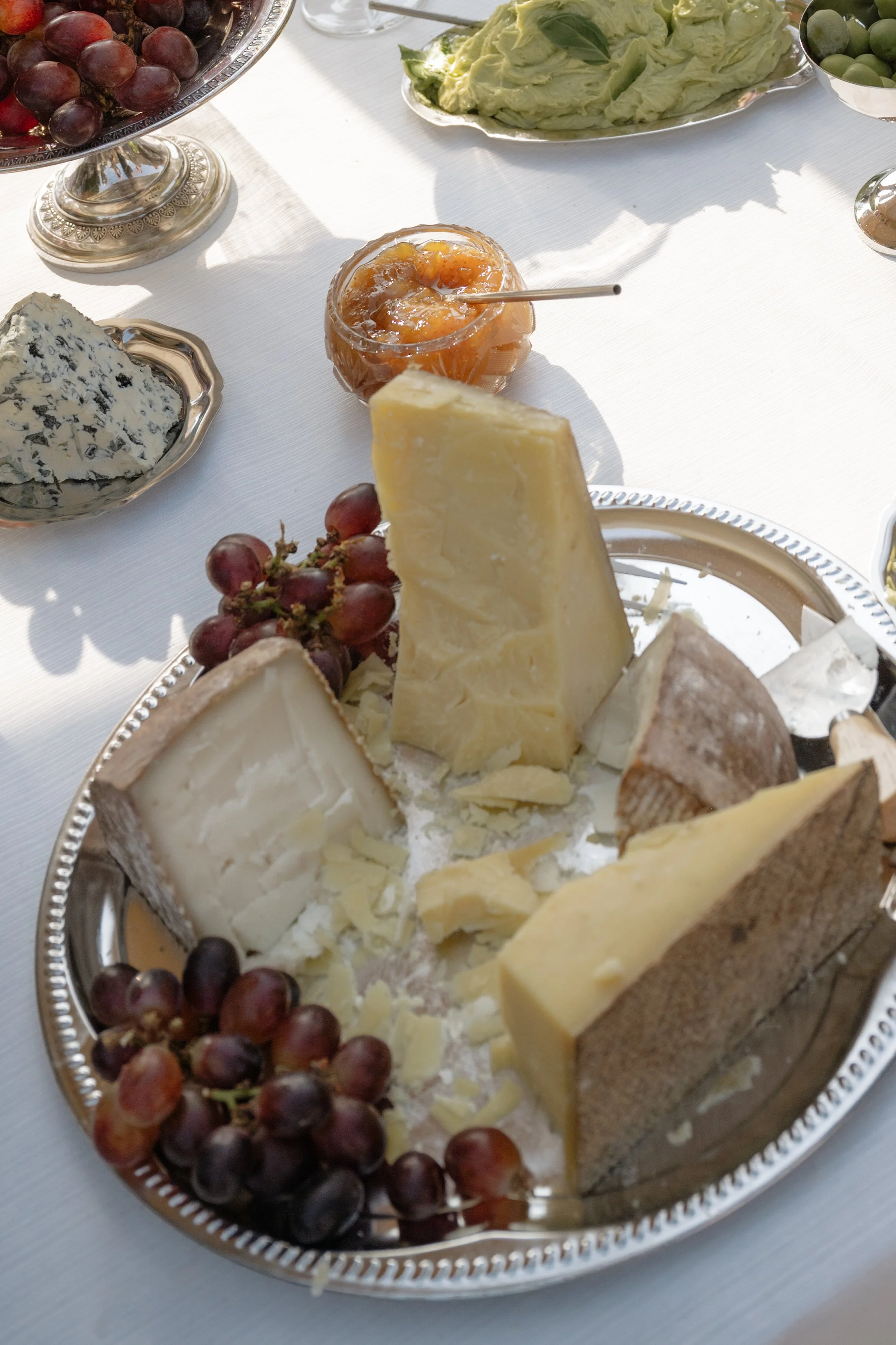 A silver platter with various cheeses, red grapes, and cheese shavings, on a white tablecloth, with additional bowls of grapes, soft cheese, and jam in the background.