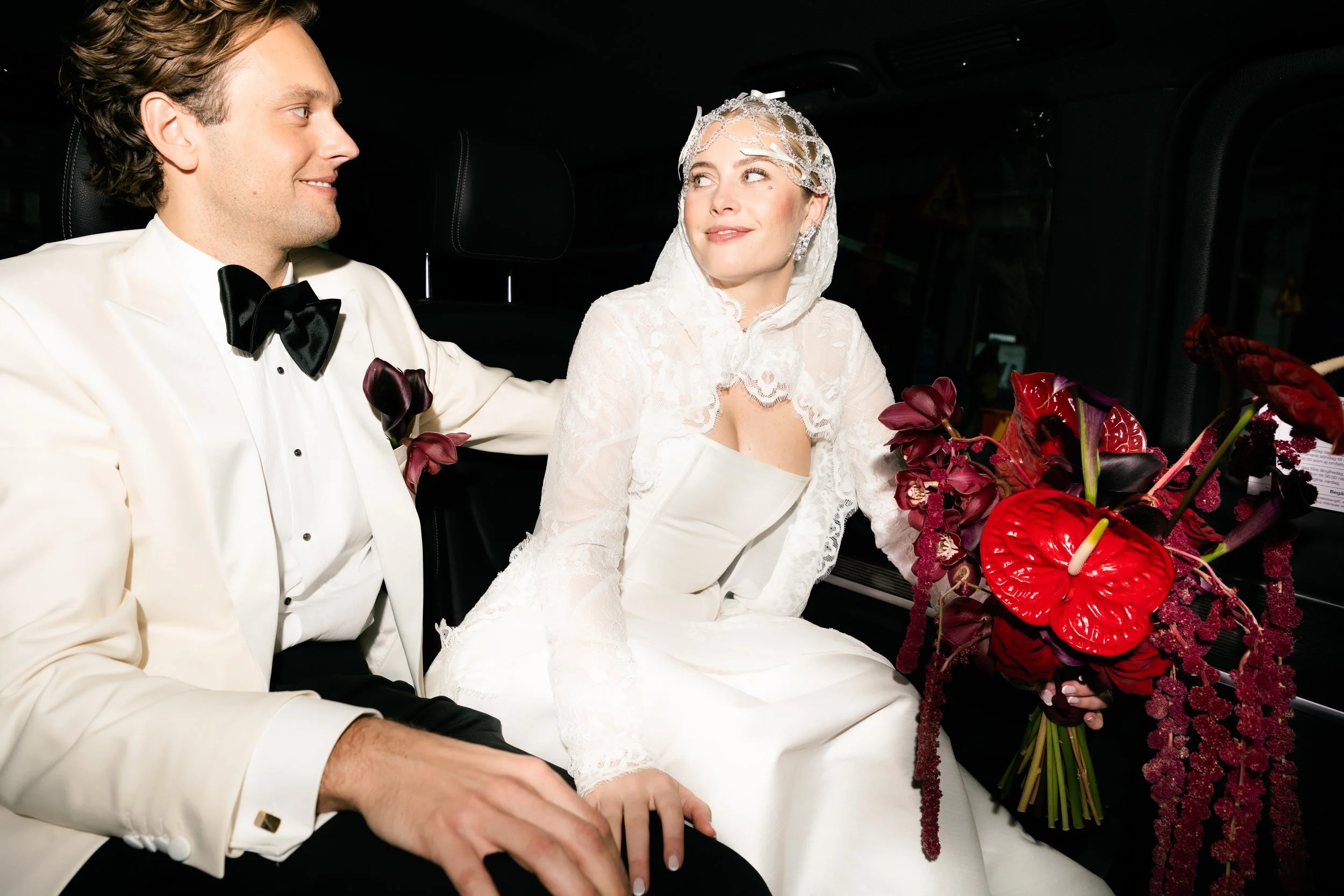 A bride and groom sitting inside a vehicle, with the groom wearing a white tuxedo and black bow tie, and the bride dressed in white with lace details, holding a bouquet of red, purple, and dark-colored flowers. They are looking at each other and smiling.