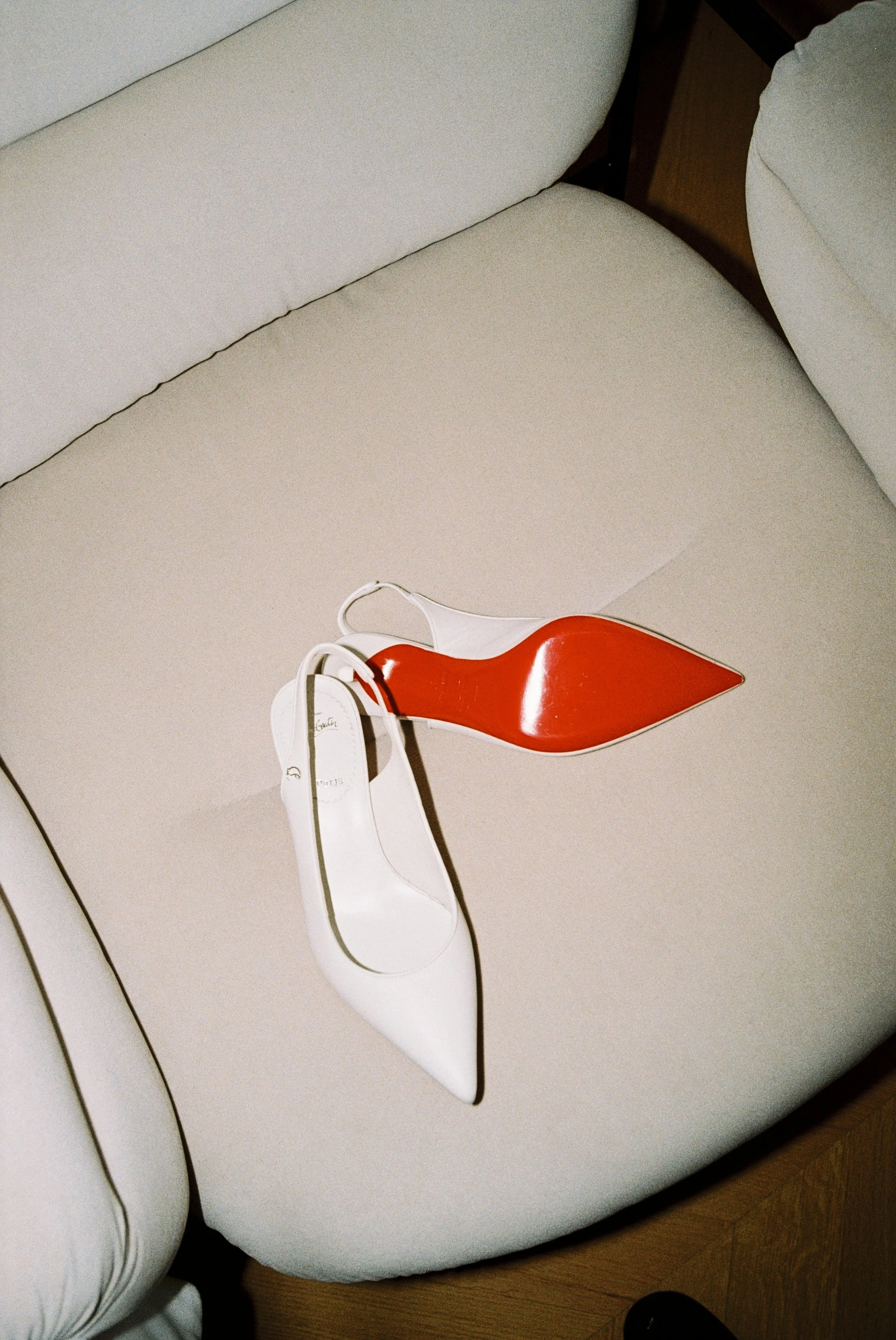 A white high-heeled shoe with a pointed toe and a red sole, placed on a beige cushioned surface.