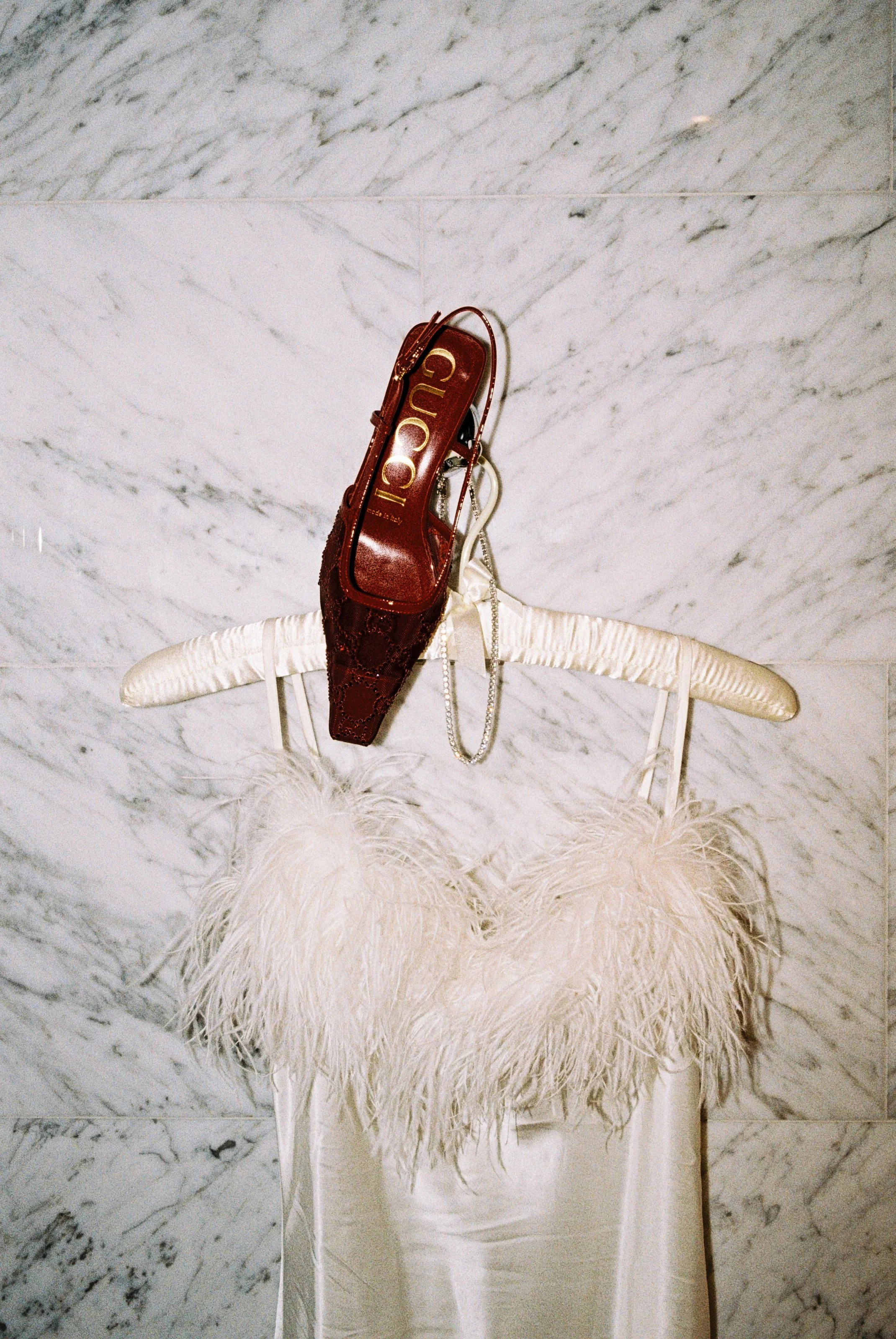 A white feathered dress hangs on a white satin-covered chair, with a white textured marble wall in the background. A red Gucci bag and a silver chain necklace hang on the hanger.