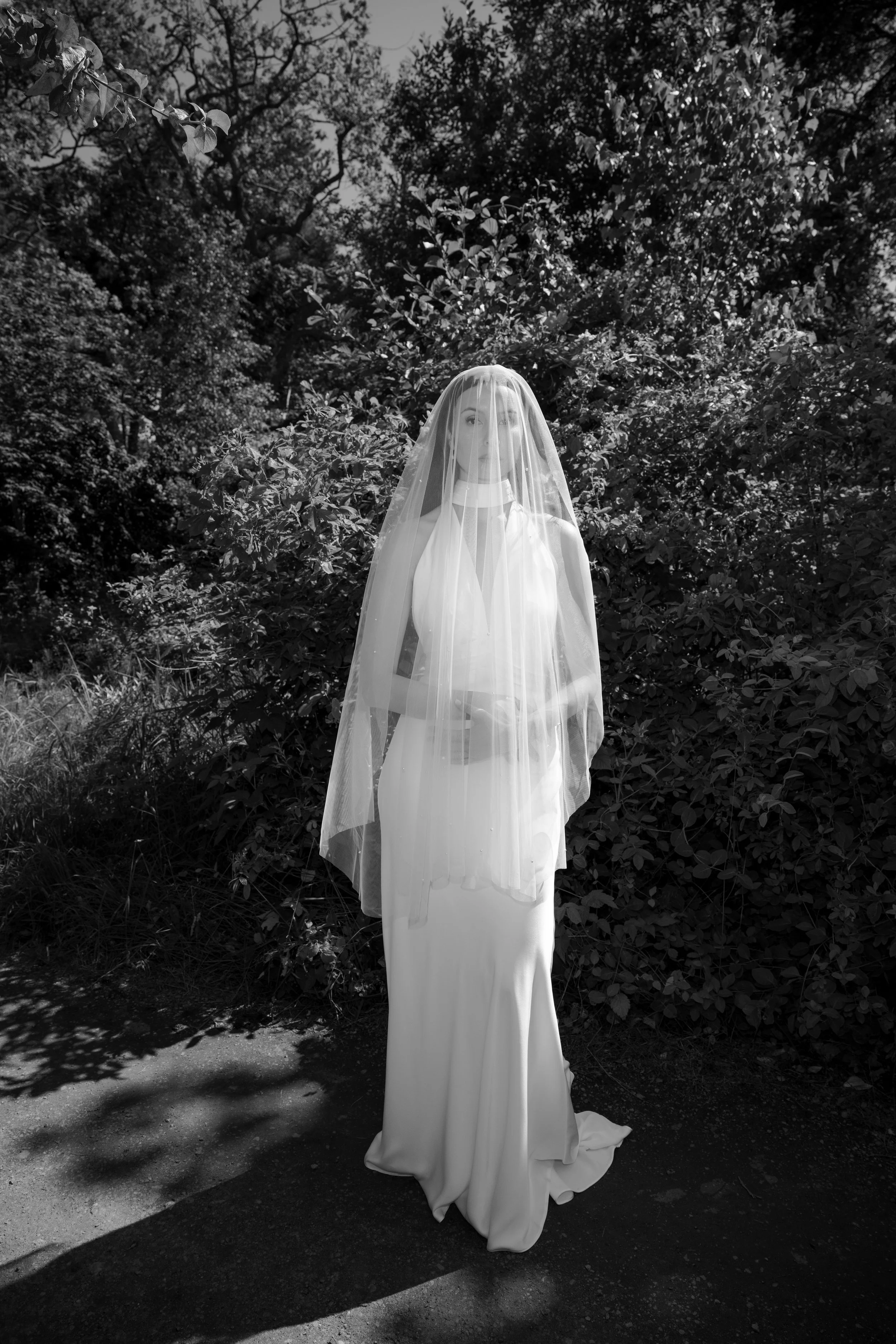 Black and white photo of a woman in a long white dress with a veil, standing outdoors in front of foliage.