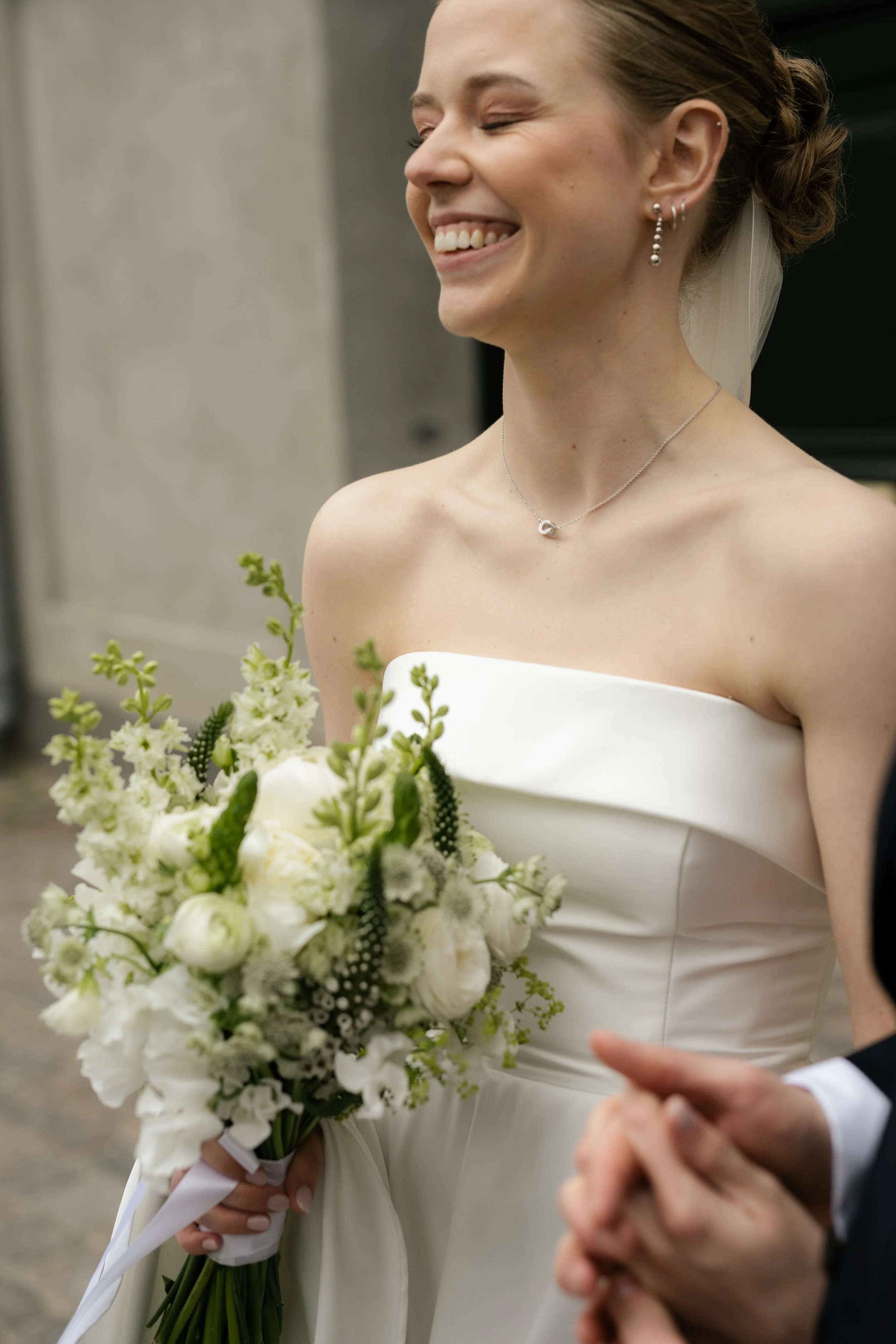 Bride in a white strapless wedding dress holding a bouquet of white flowers, smiling with eyes closed, standing outdoors.