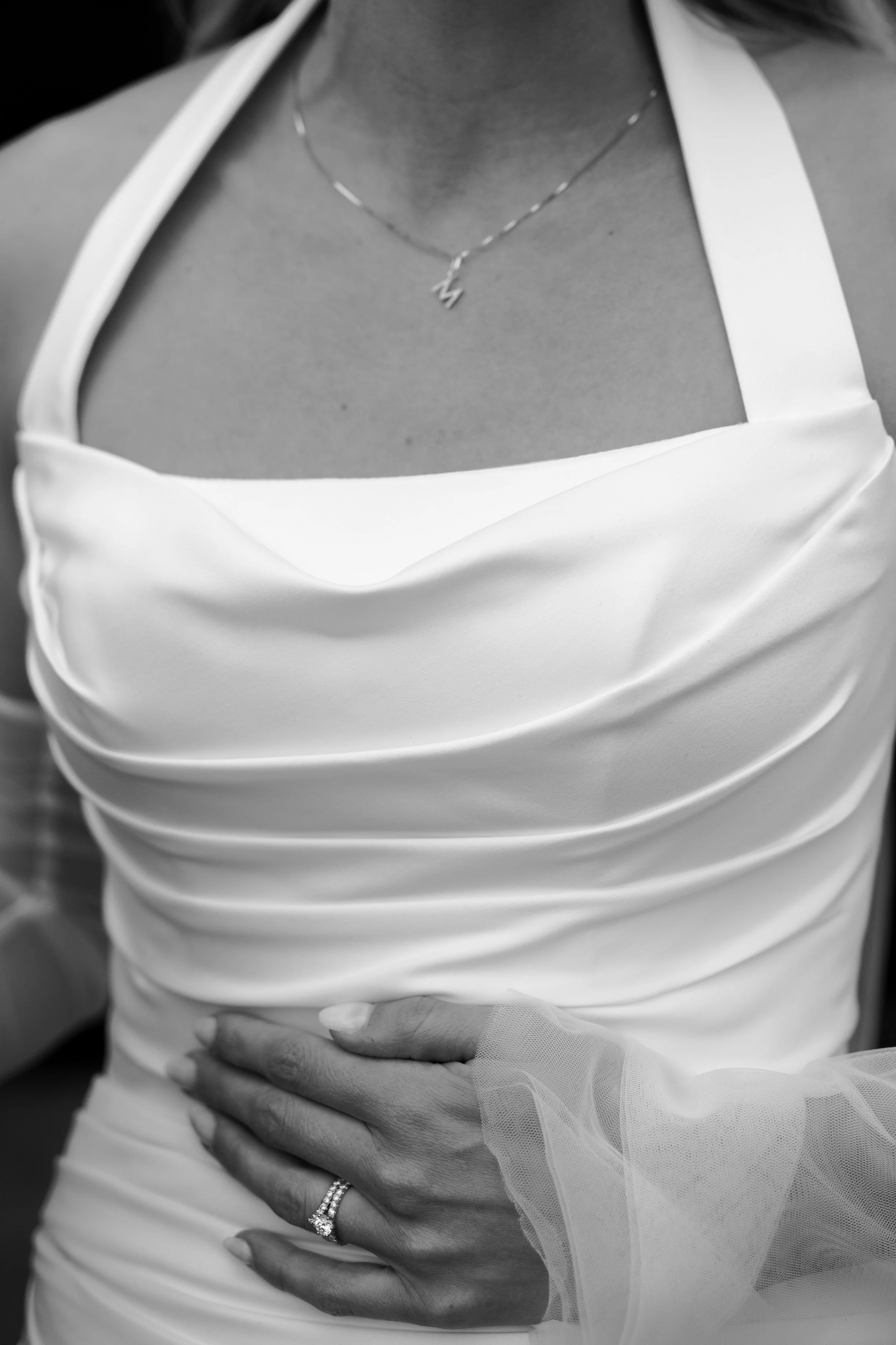 Close-up of a woman in a wedding dress with a hand on her waist, wearing a ring and a delicate necklace with a lightning bolt pendant, in black and white.