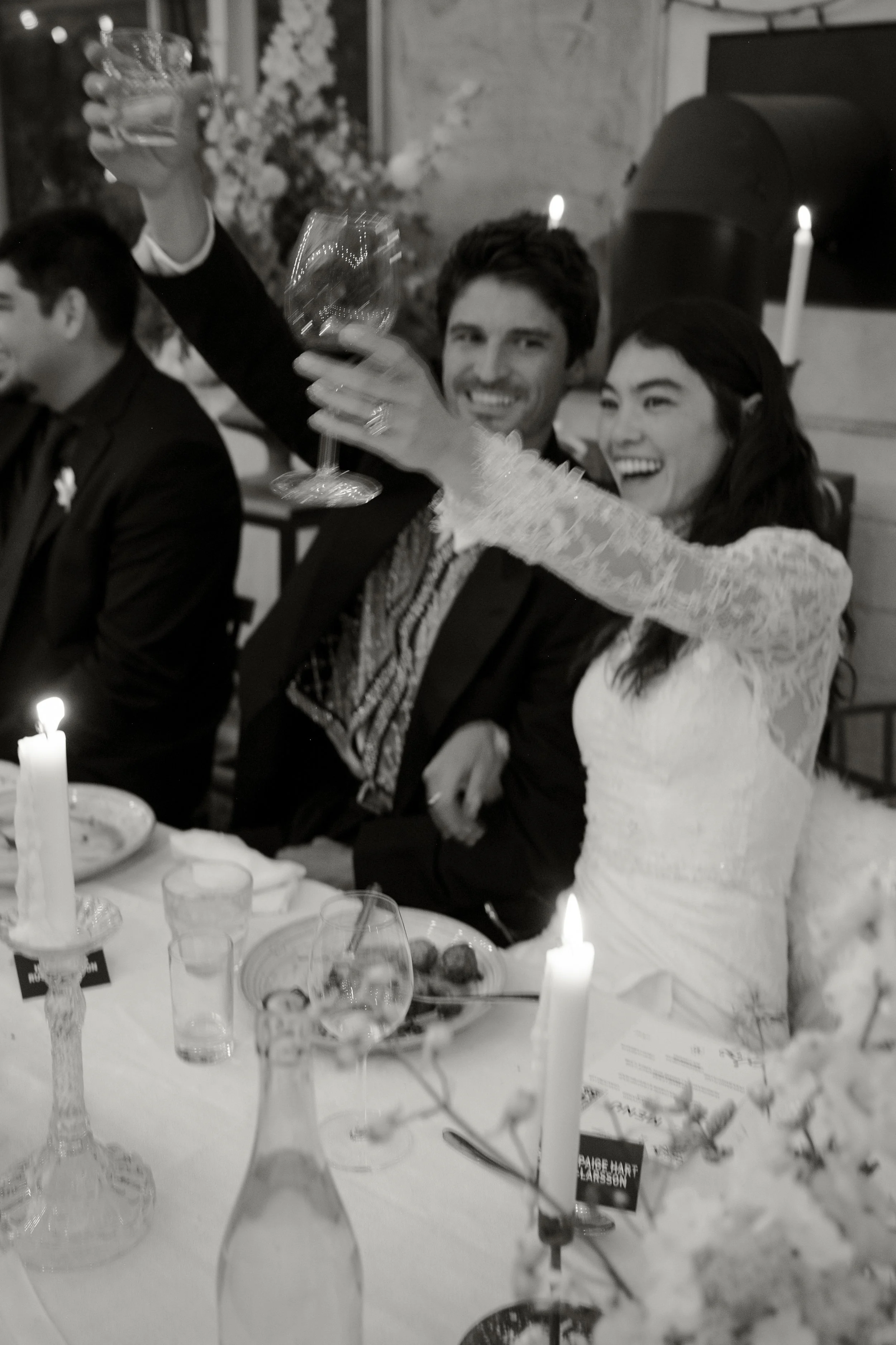 A black and white photo of a joyful celebration at a wedding reception, with a bride and groom raising glasses in a toast, surrounded by friends and family seated at a decorated table.