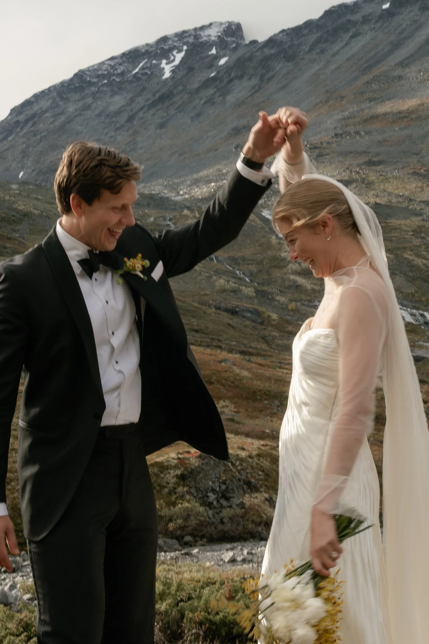 A newly married couple dancing outdoors with a mountainous landscape in the background, the groom in a tuxedo and the bride in a white wedding dress holding a bouquet.