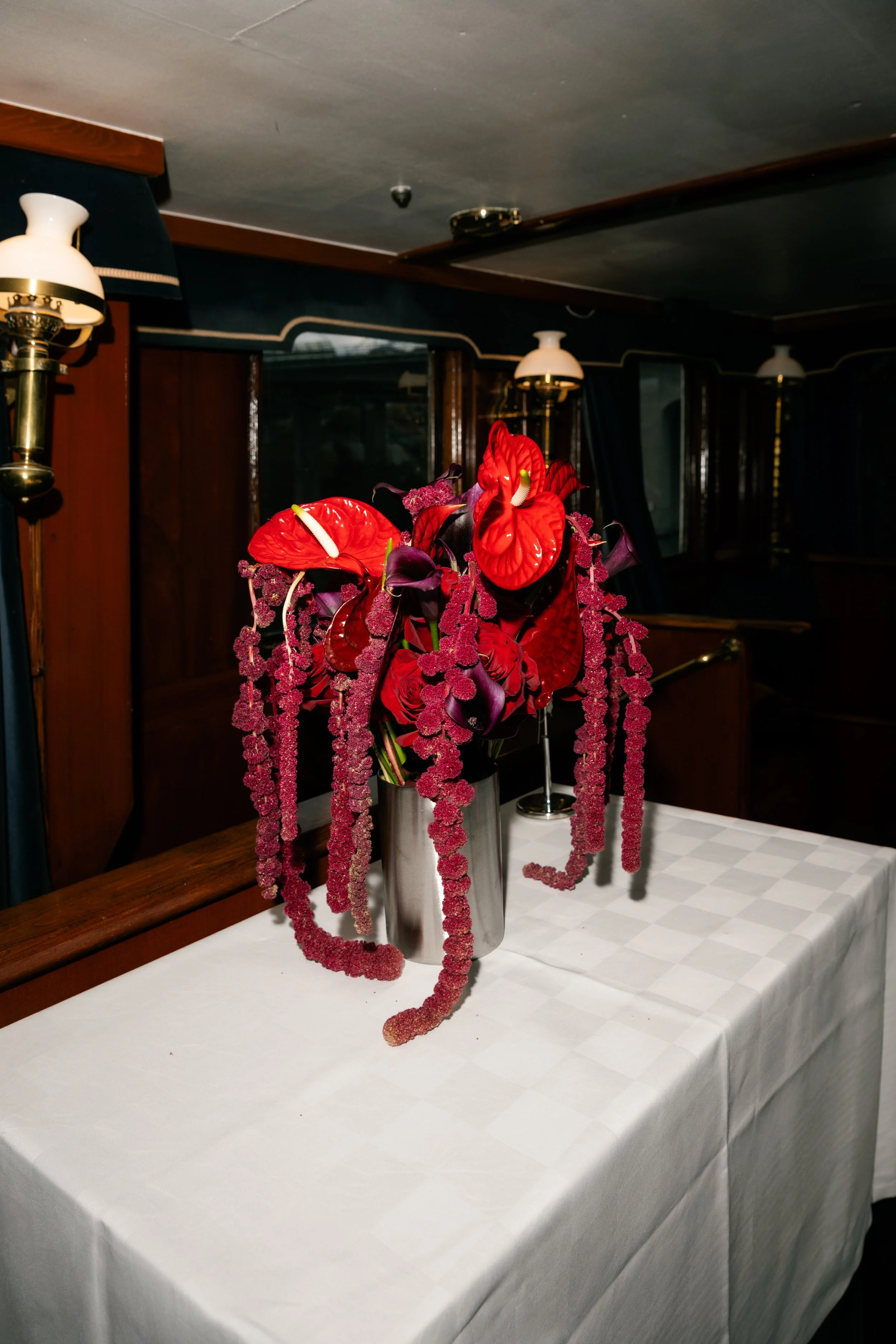 A floral arrangement with red and purple flowers in a silver vase on a white tablecloth table, with two lamps and wood-paneled walls in the background.