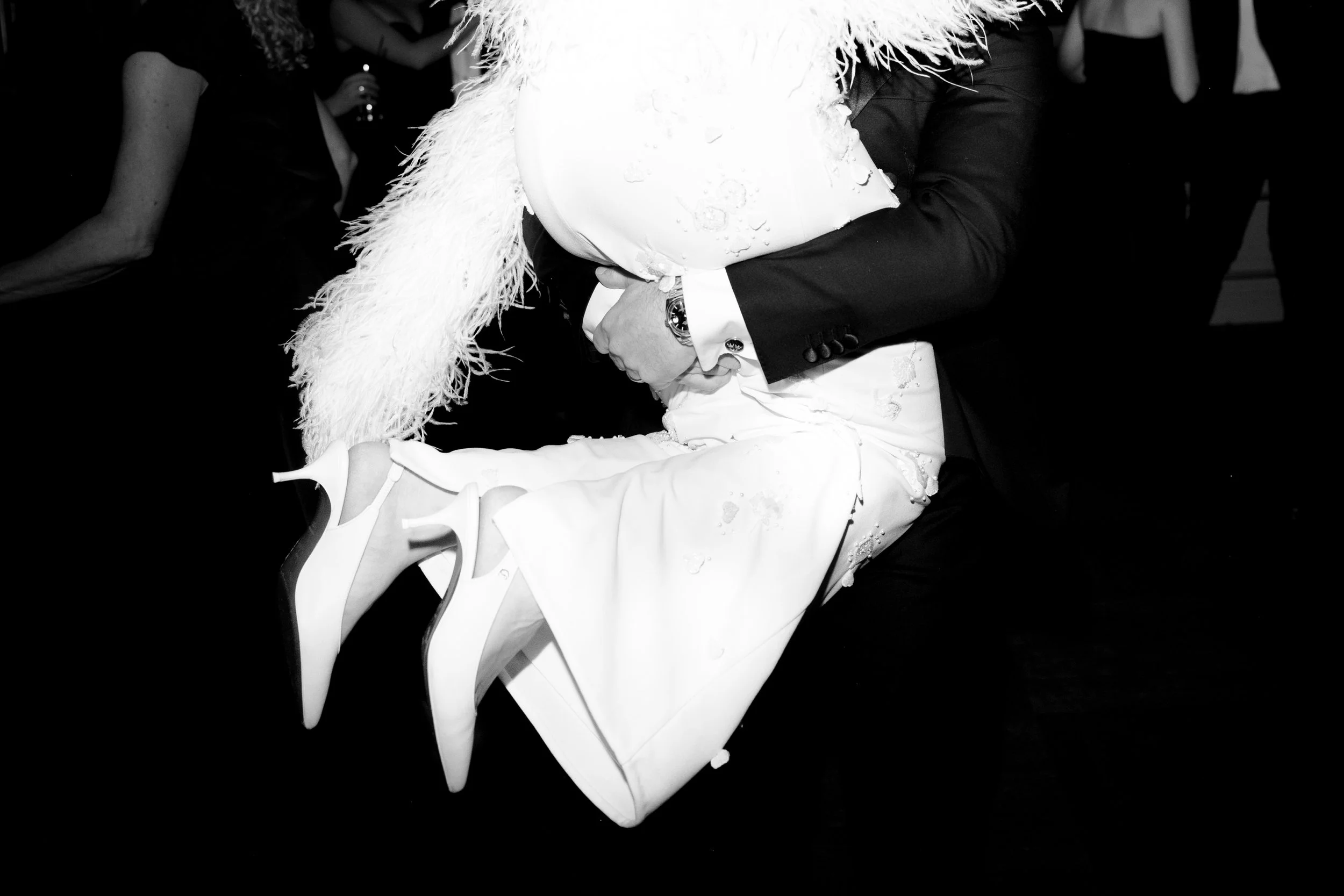 A person in a tuxedo is holding another person who is wearing a long dress and white high heels, in a dance pose at a formal event.