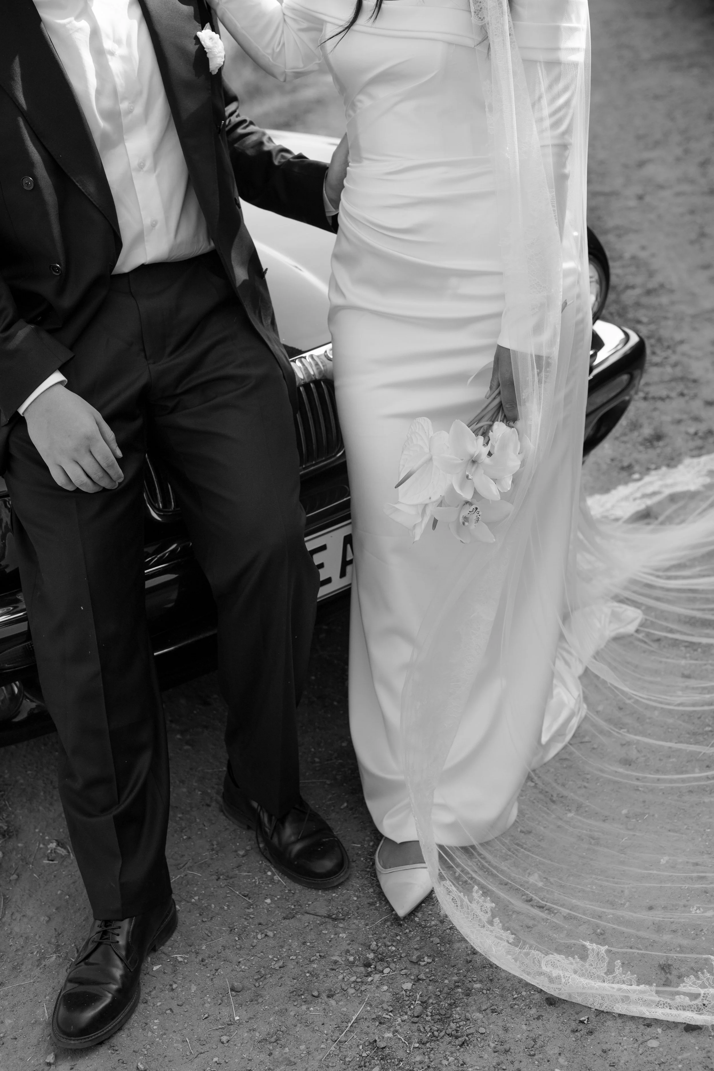  Bride and groom standing by a car, with bride holding a bouquet of flowers. Black and white photo.